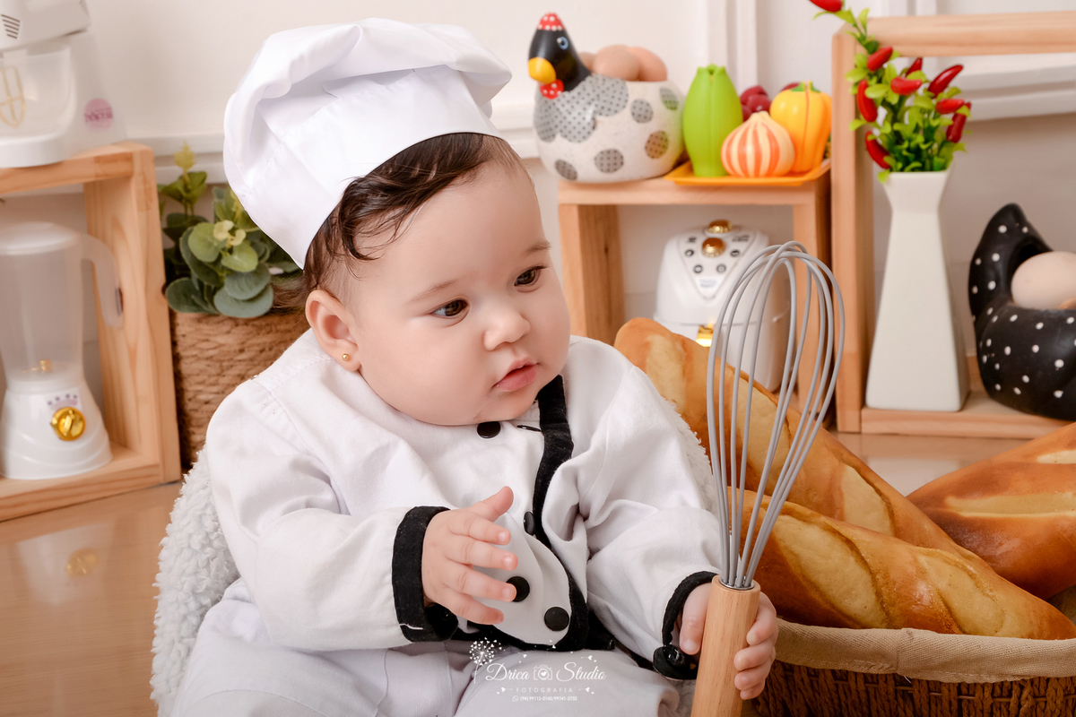 drica studio - ensaio fotográfico infantil - acompanhamento - bebê vestido de chef de cozinha, sentando em poltrona segurando um batedor de massa, ao seu lado um cesto de pães - galinha decorativa - legumes de brinquedo-fundo branco com boiserie