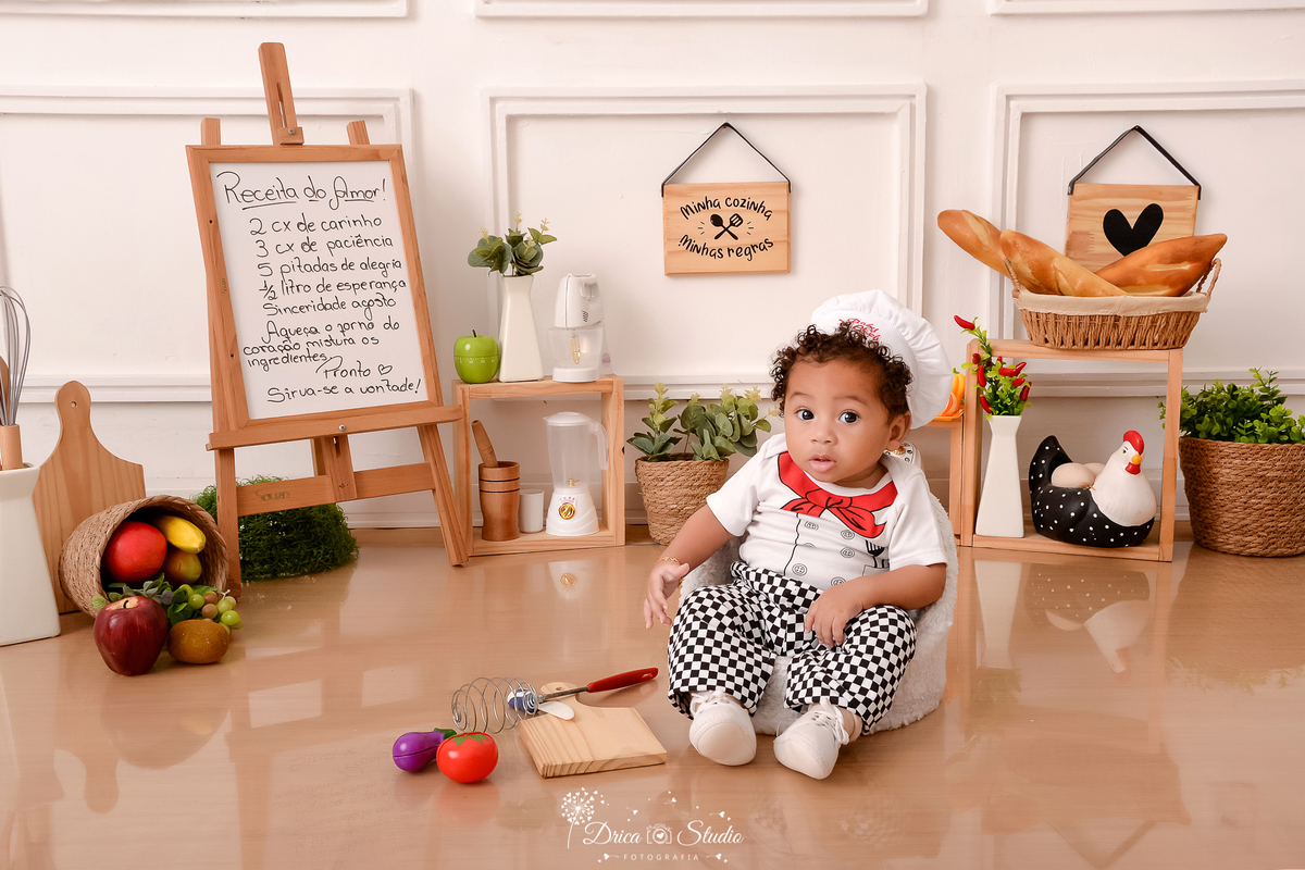 drica studio - ensaio fotográfico infantil - acompanhamento - bebê sentado em poltrona, vestido de chef de cozinha - cesto com frutas - placa com receita - utensílios de cozinha - cesto com arbusto - cesto com pães - piso amadeiradodrica studio - ensaio f