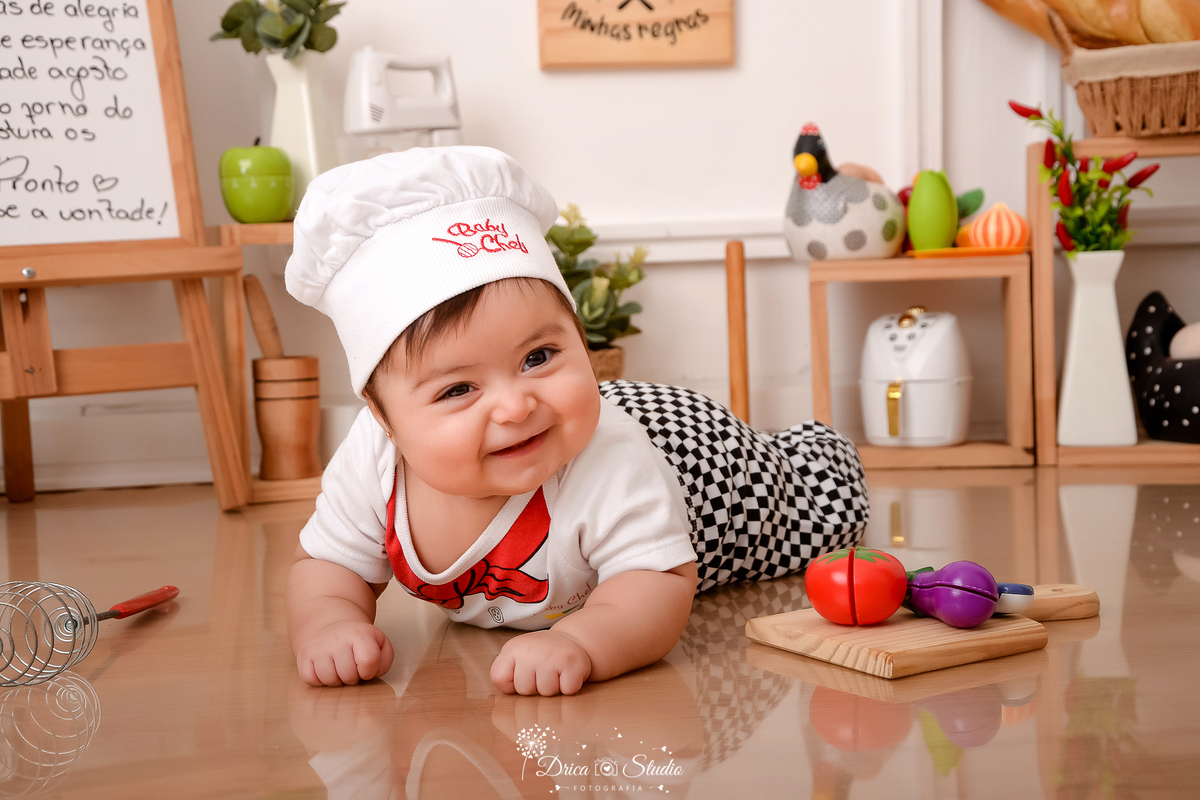 drica studio - ensaio fotográfico infantil - acompanhamento - bebê deitada de bruços no chão, vestinda com roupa de chef de cozinha - tomate e beringela de brinquedo - panela eletrica - galinha e ovos decorativos - vaso branco com pimenteira - pilão 