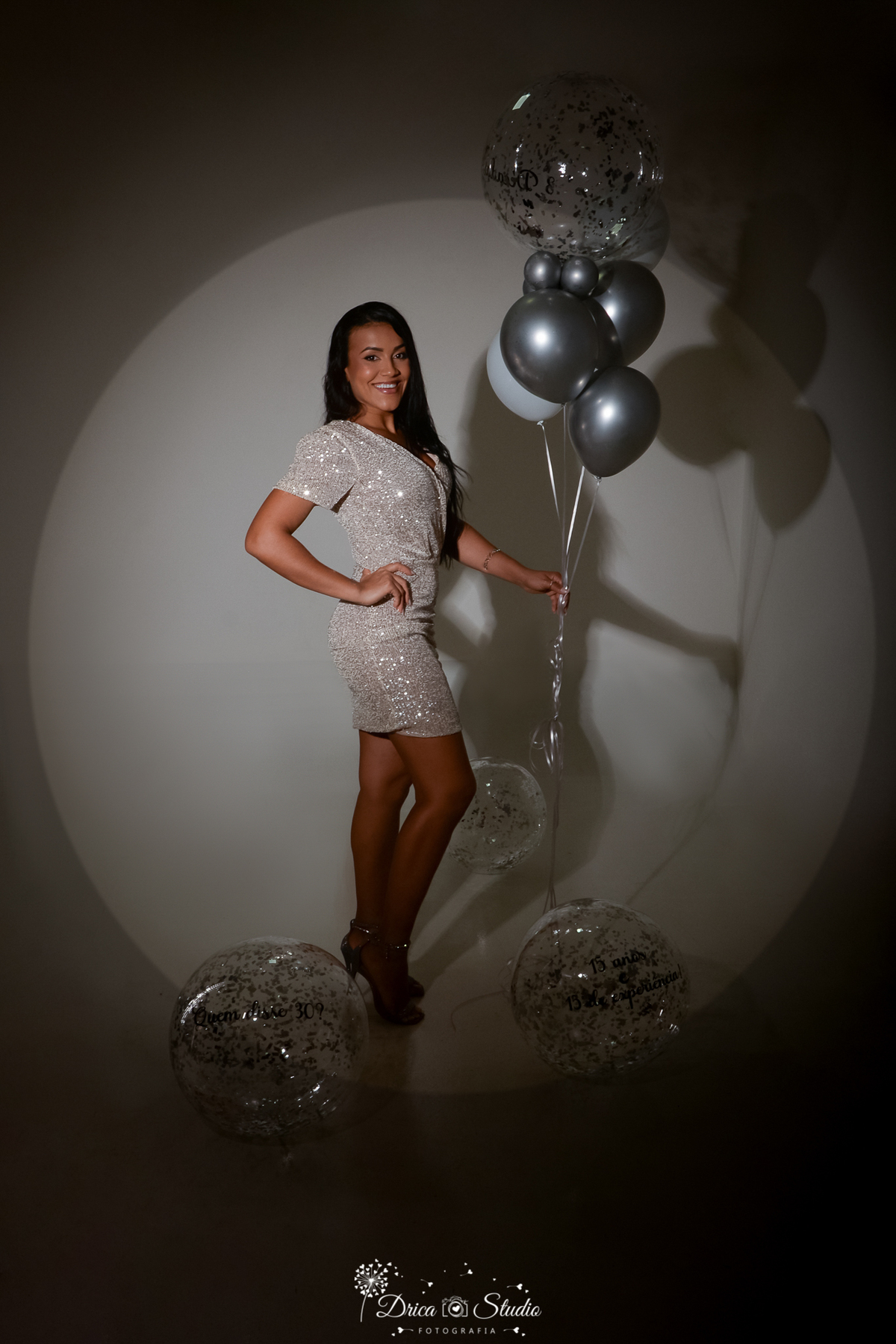 drica studio - ensaio fotográfico - xinguara - mulher usa um vestido curto todo em brilho, segurando um conjuto de balões prateados, transparente e com glitter. A iluminação cria um círculo de luz suave ao fundo, mostrando sua silhueta
