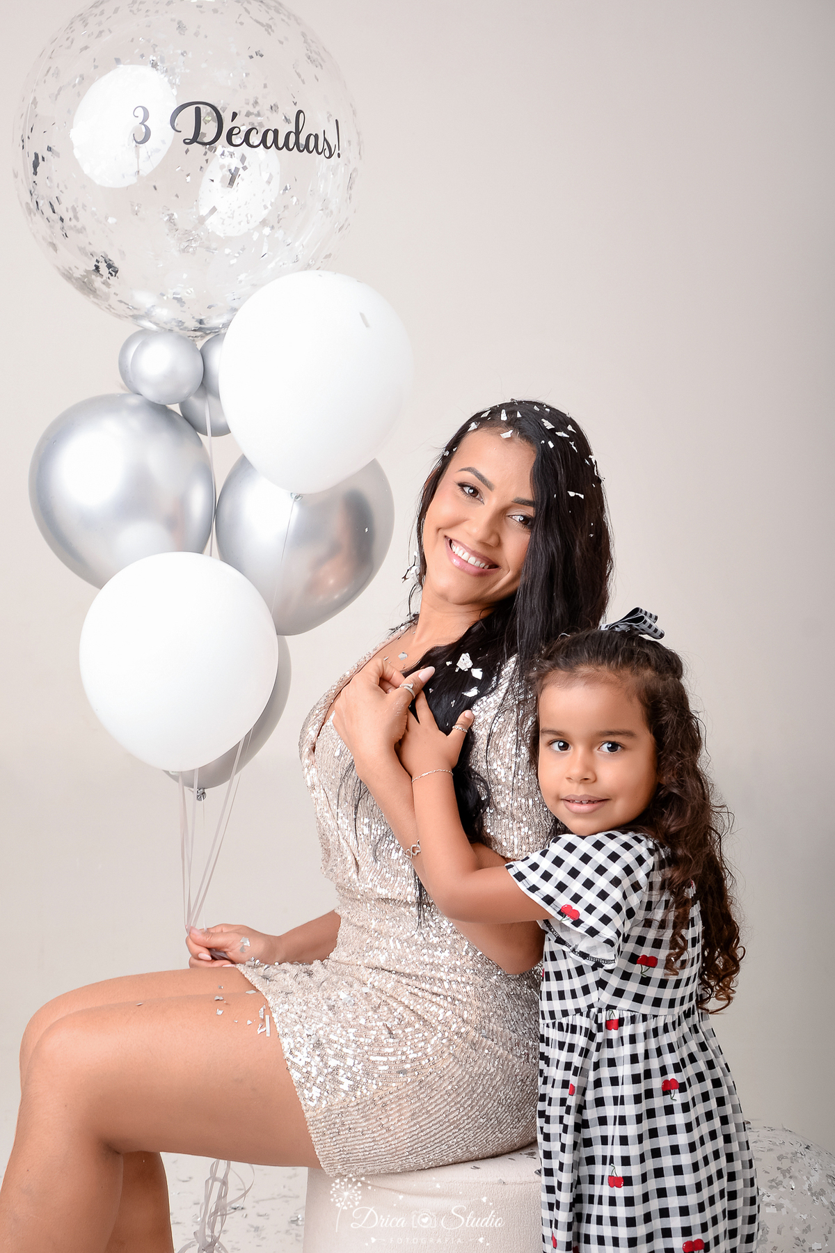 dirca studio - ensaio fotografico - xinguara - filha abraça a mãe - mulher sentada, usando vestido curto, segurando balões, usa um vestido curto brilhante, em tom champagne - Ao seu lado, uma criança a abraça, apoiando-se nela, menina usa vestido xadrez.