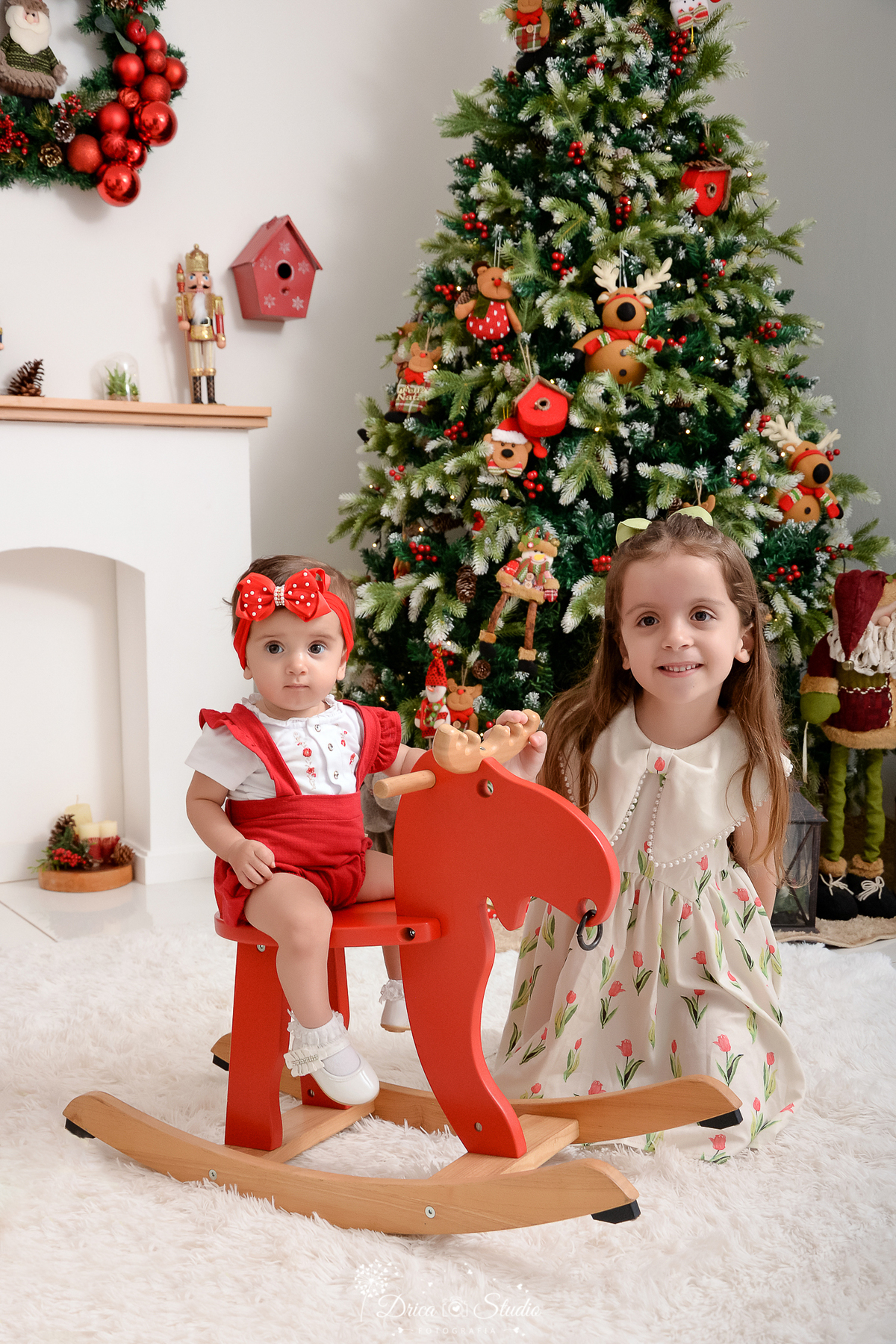 drica studio - ensaio fotografico - xinguara - fotos de natal - bebê sentada em rena vermelha de madeira e irmã mais velha sentada ao lado - a bebê usa macaquinho vermelho com blusa branca e a menina bestido bege com flores bordadas - árvore de natal 