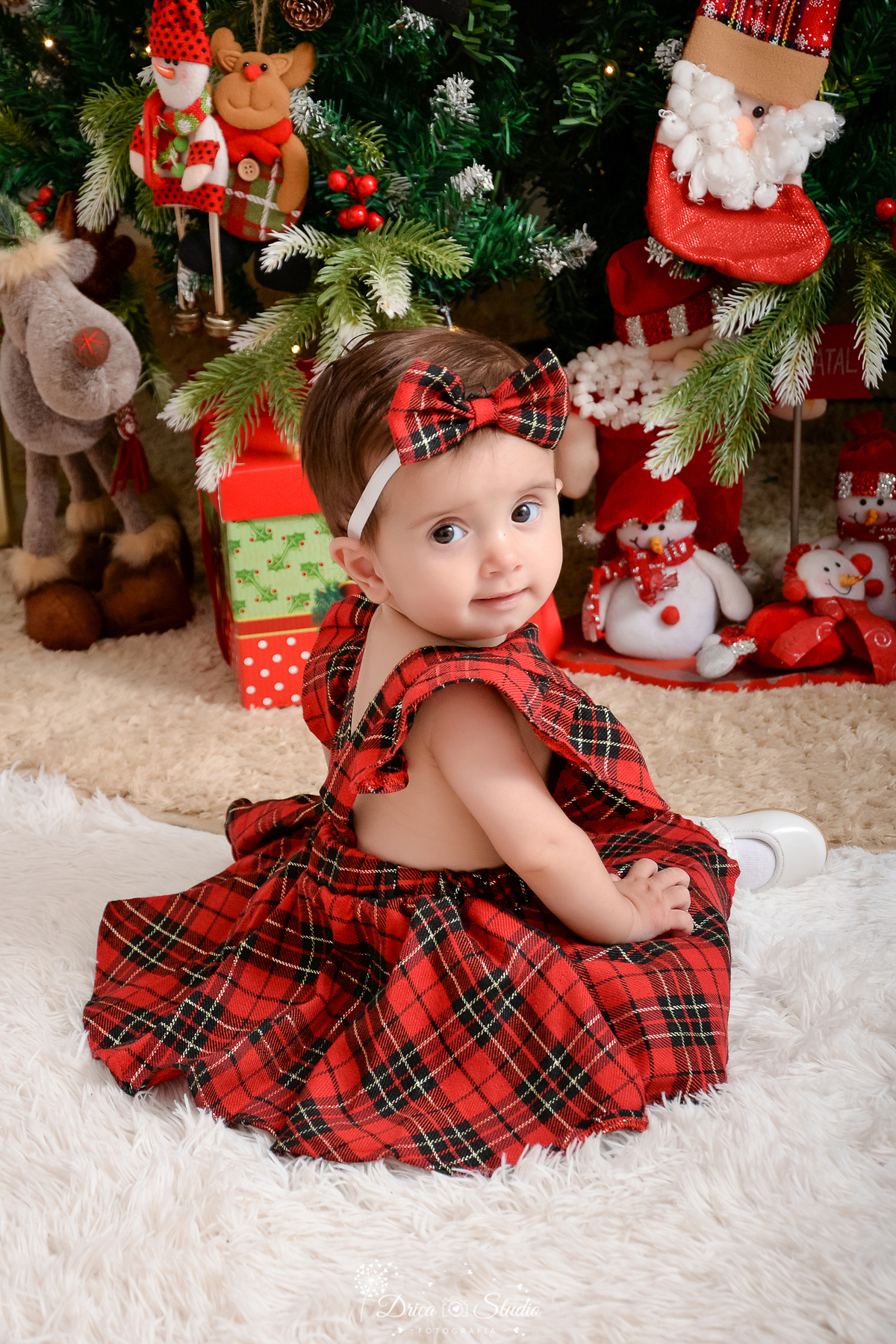 drica studio - ensaio fotografico - xinguara - fotos de natal - bebê vestinda com jardineira xadres vermelho com preto, sentada em carpete branco - bonecos de neve de pelúcia - caixas de presentes - rena de pelúcia - laço xadrez vermelho com preto