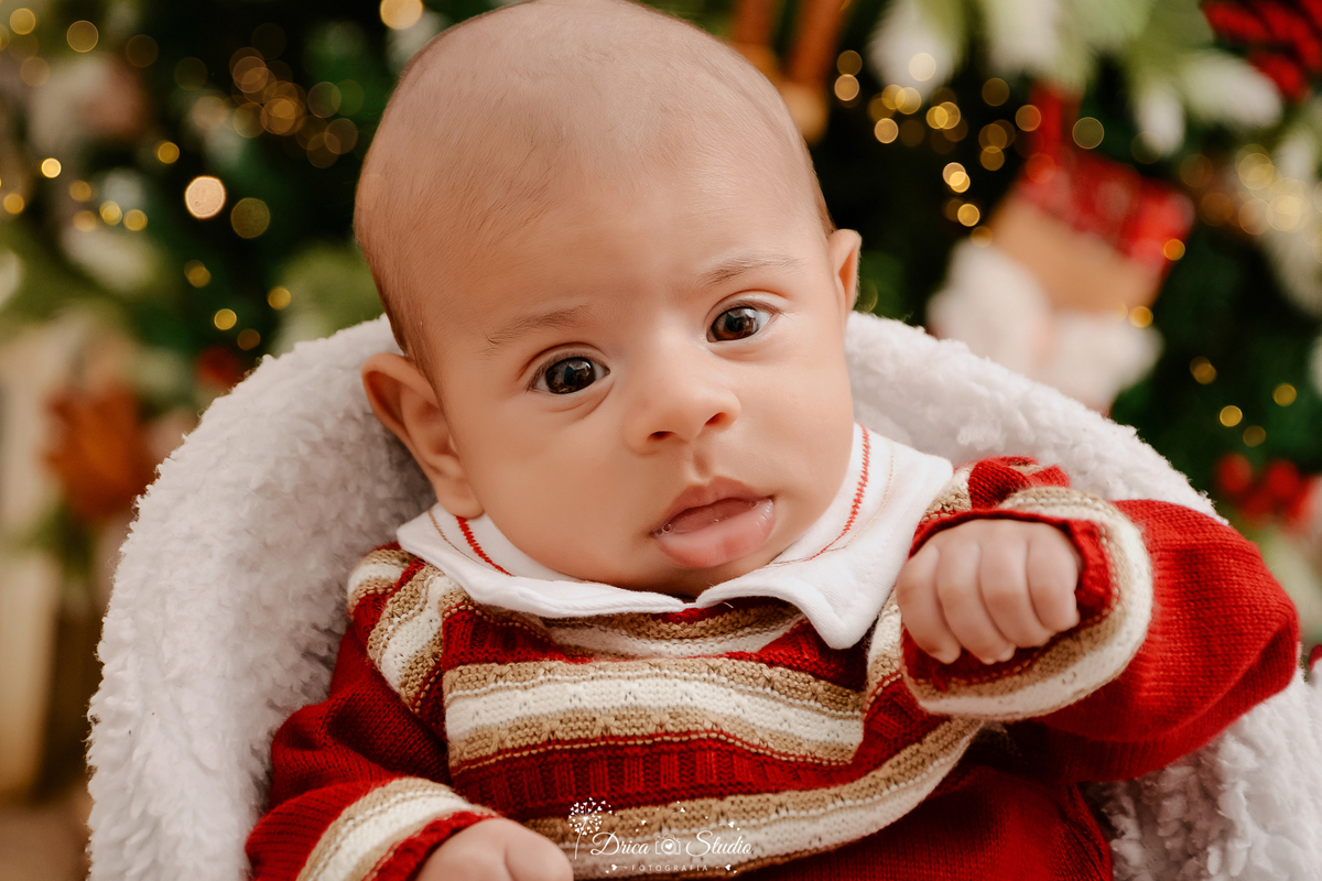 drica studio - ensaio fotografico - xinguara - fotos de natal - bebê vestindo macaquinho vermelho com listras brancas, sentado em poltrona branca - árvore de natal com piscas