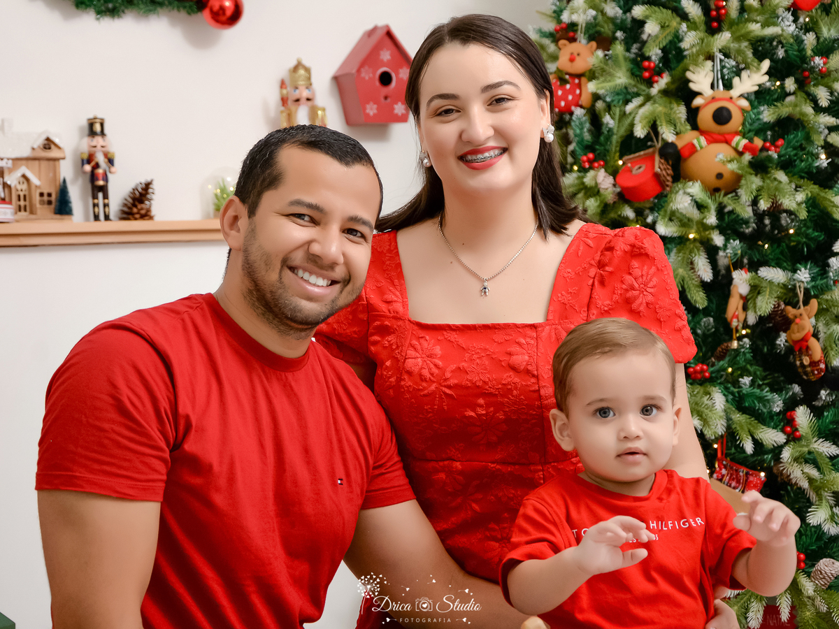 drica studio - ensaio fotografico - xinguara - fotos de natal - homem vestindo camisa vermelha - mulher vestindo vestido vermelho e bebê usando camisa vermelha - árvore de natal - quebra nozes - casinha de enfeite - fundo branco