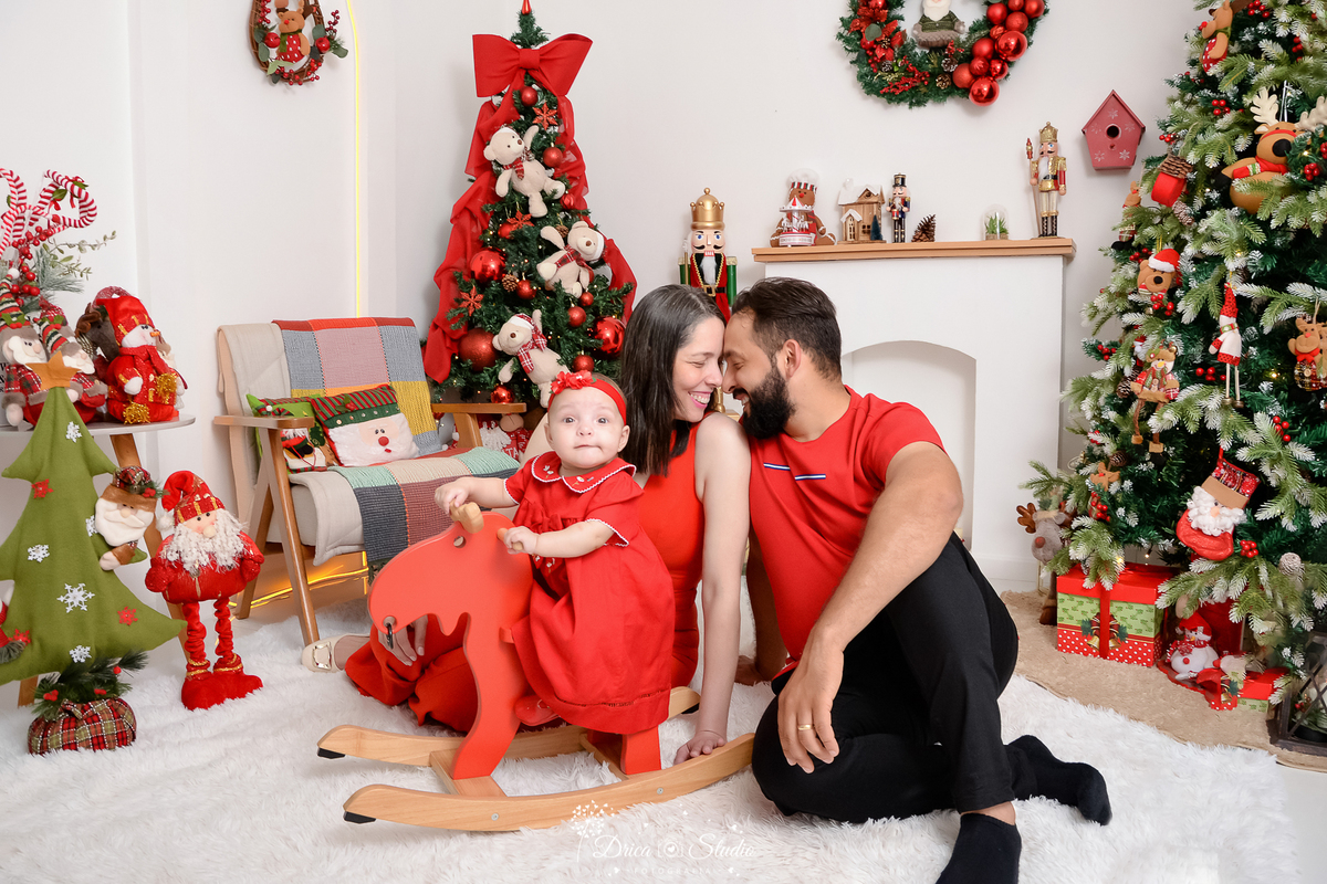 drica studio - ensaio fotografico - xinguara - fotos de natal - casal sentando no chão com rosco levenente encostado - homem veste camisa vervelha e calça preta - mulher e bebê veste vestido vermelho - árvore de natal - papai noel de pelúcia -fundo branco