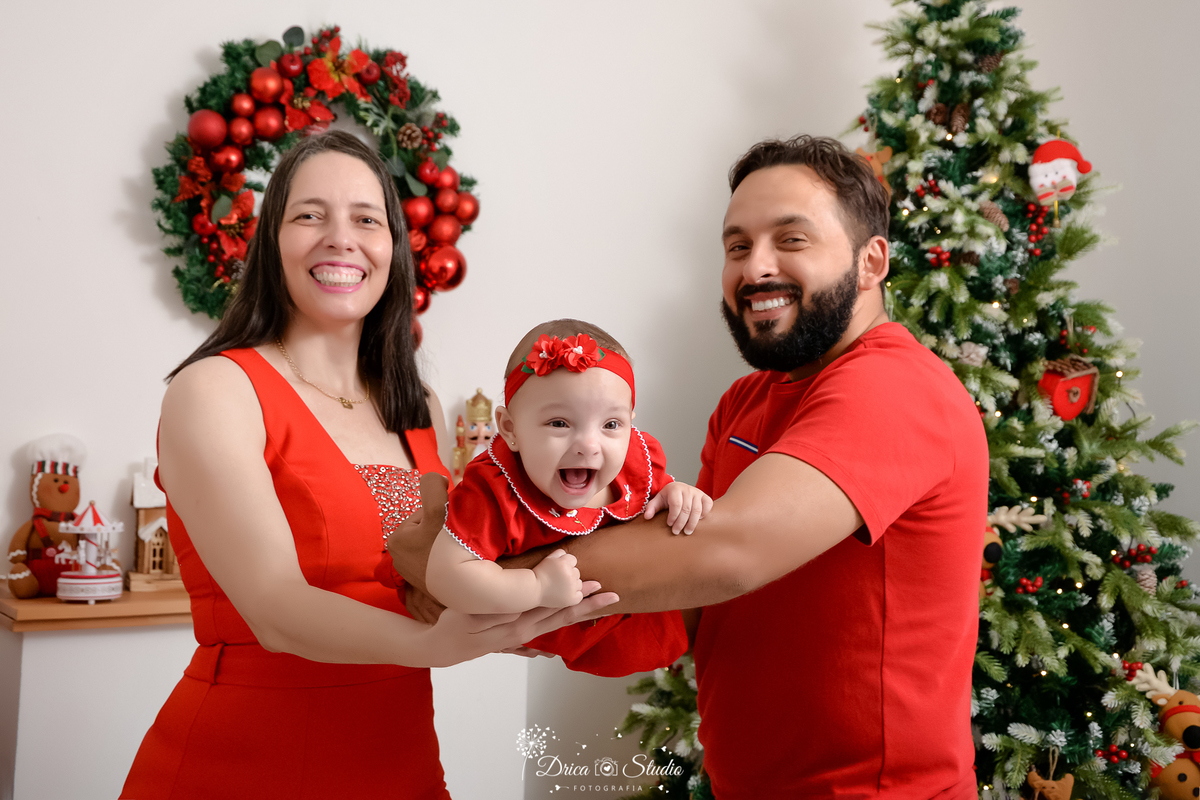 drica studio - ensaio fotografico - xinguara - fotos de natal -  pais e a bebê vestindo roupas vermelha - o casal sustentam a filha nos braços - tiara vermelha - árvore de natal - guirlanda - lareira branca - caixinha de música em forma e carrocel 