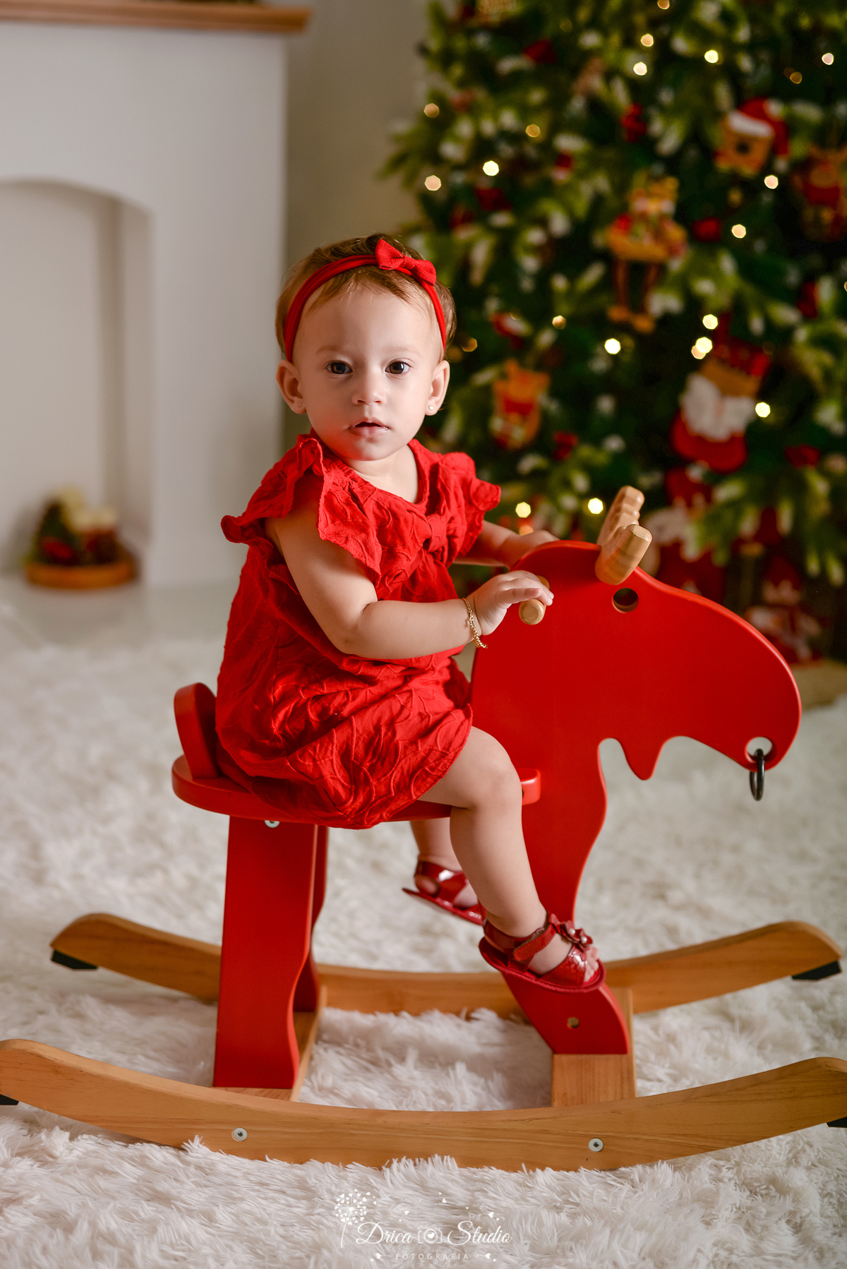 drica studio - ensaio fotografico - xinguara - fotos de natal - árvore de natal - rena vermelha de madeira - bebê usando vestido vermelho - carpete felpudo branco - lareira branca
