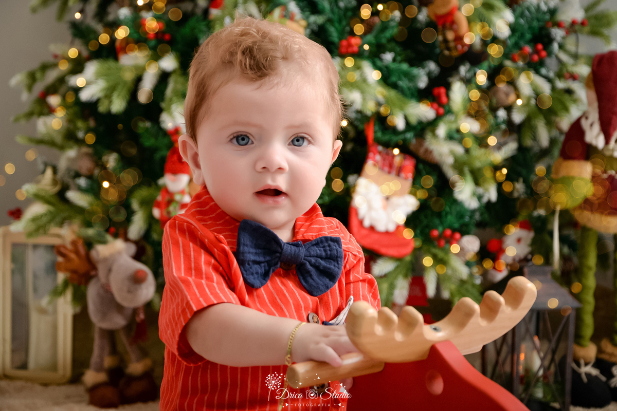 drica studio - ensaio fotografico - xinguara - fotos de natal - bebê usando camisa vermelha com listras brancas e gravata borboleta azul - árvore de natal - rena de pelúcia - lampião com vela - enfeite de natal