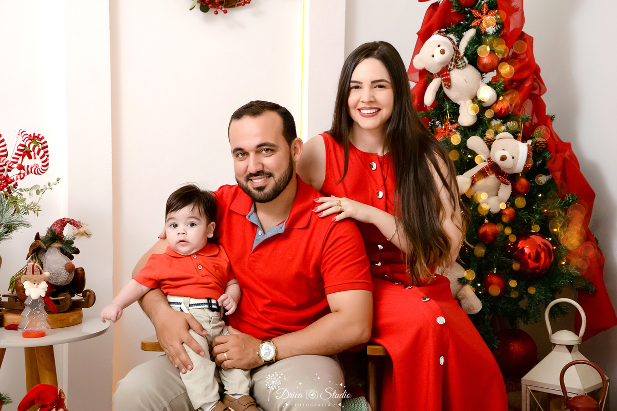 drica studio - ensaio fotografico - xinguara - fotos de natal - pai sentado em poltrona, esposa sentado no braça da poltrona e bebê sentado no colo do pai, familia usa tom vermelho e bege claro - árvore de natal - ursinhos natalino - enfeites de natal