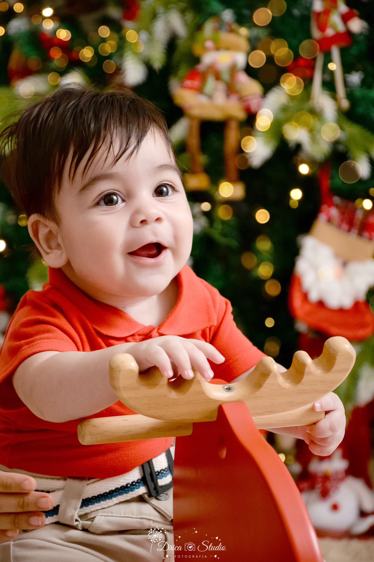 drica studio - ensaio fotografico - xinguara - fotos de natal - bebê usando camisa vermelha e calça bege, sentado em rena vermelha de madeira - árvore de natal - boneco de neve de pelúcia