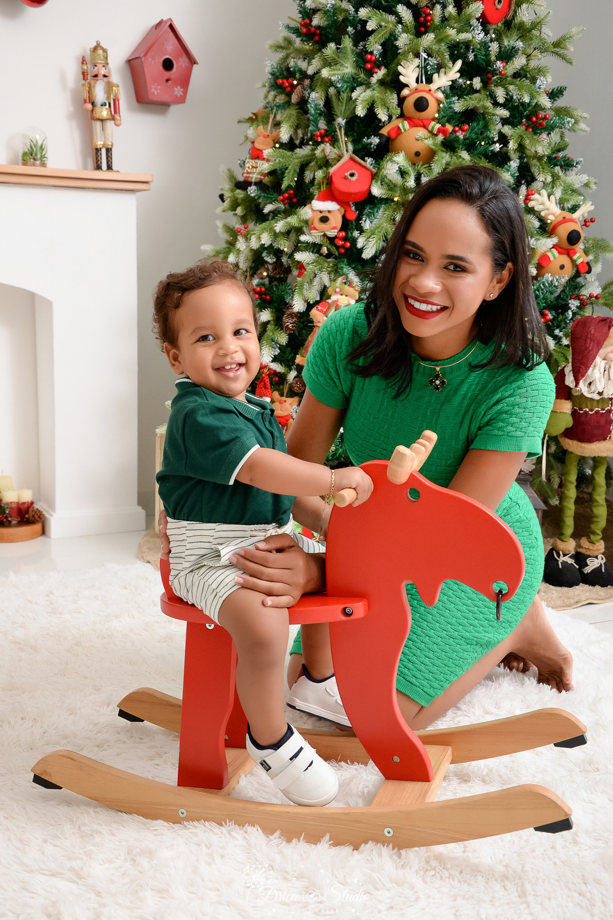 drica studio - ensaio fotografico - xinguara - fotos de natal - mãe sentada sobre as perna segurando bebê sentado em rena de madeira - vestido longo verde - camisa gola polo verde escuro e short bege - árvore de natal - papai noel - lareira banca-