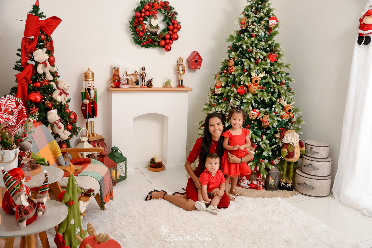 drica studio - ensaio fotografico - xinguara - fotos de natal - mâe com casal de filhos pequenos, ambos vestem roupas vermelha - árvore grande de natal - lareira branca - quebra nozes - guirlanda - papai noel de pelúcia - fundo branco - fundo branco