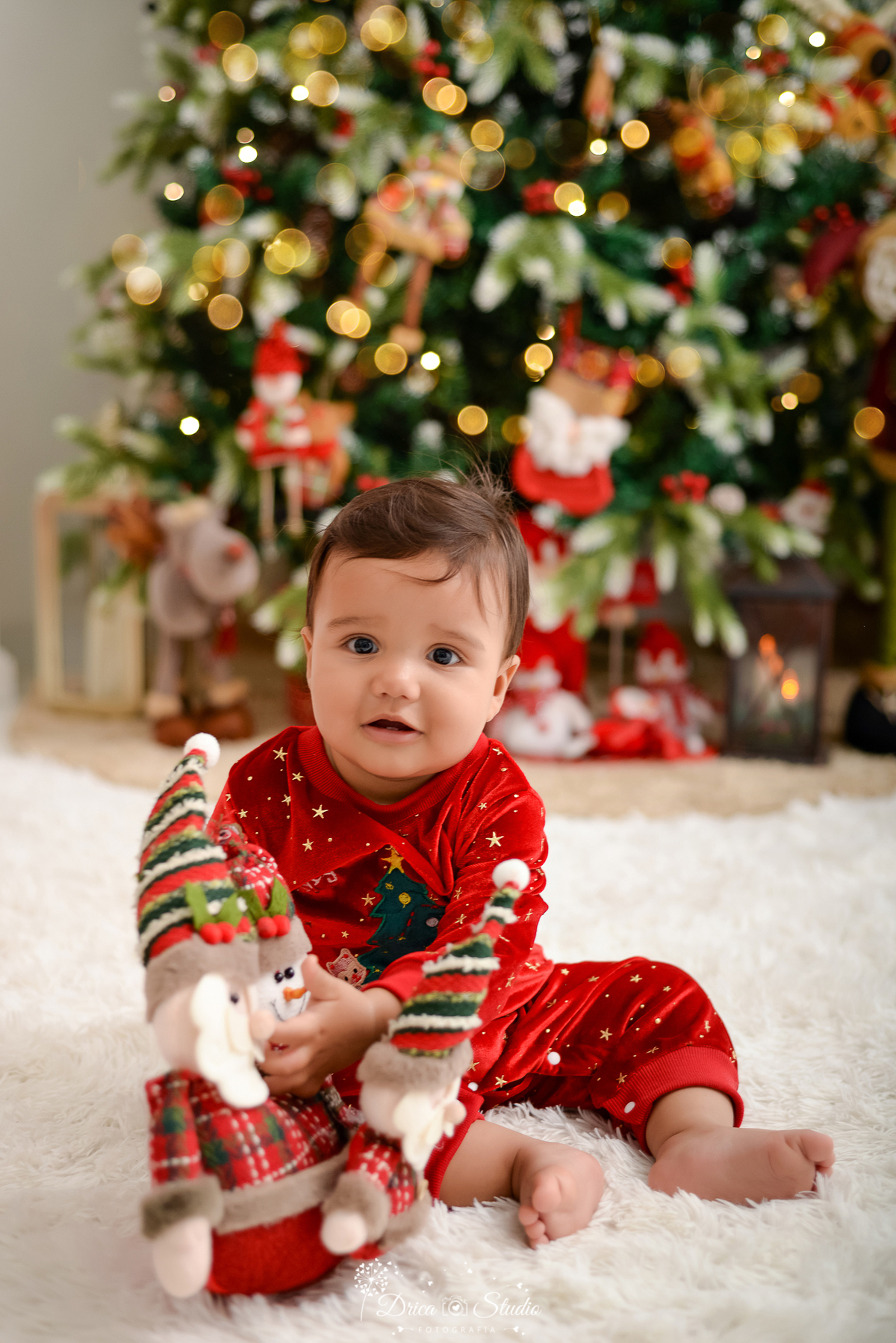 drica studio - ensaio fotografico - xinguara - fotos de natal - bebê sentado no chão segurando papai noel de pelucia, vestido em macaquinho de veludo vermelho - ao fundo árvore de natal - rena de pelucia - carpete branco