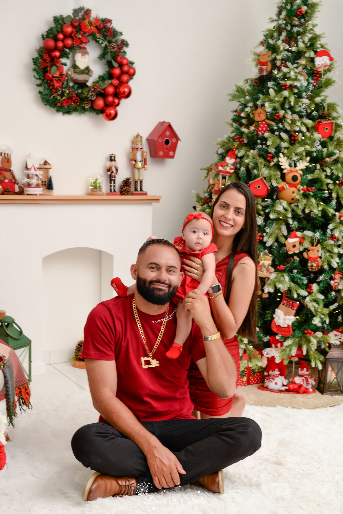 drica studio - ensaio fotografico - xinguara - fotos de natal - pai sentado no chão e mãe segurando bebê sentada nos ombros do pai - vertidos vermelho e camisa vermelha - árvore de natal - lareira branca - quebra nozes - fundo branco