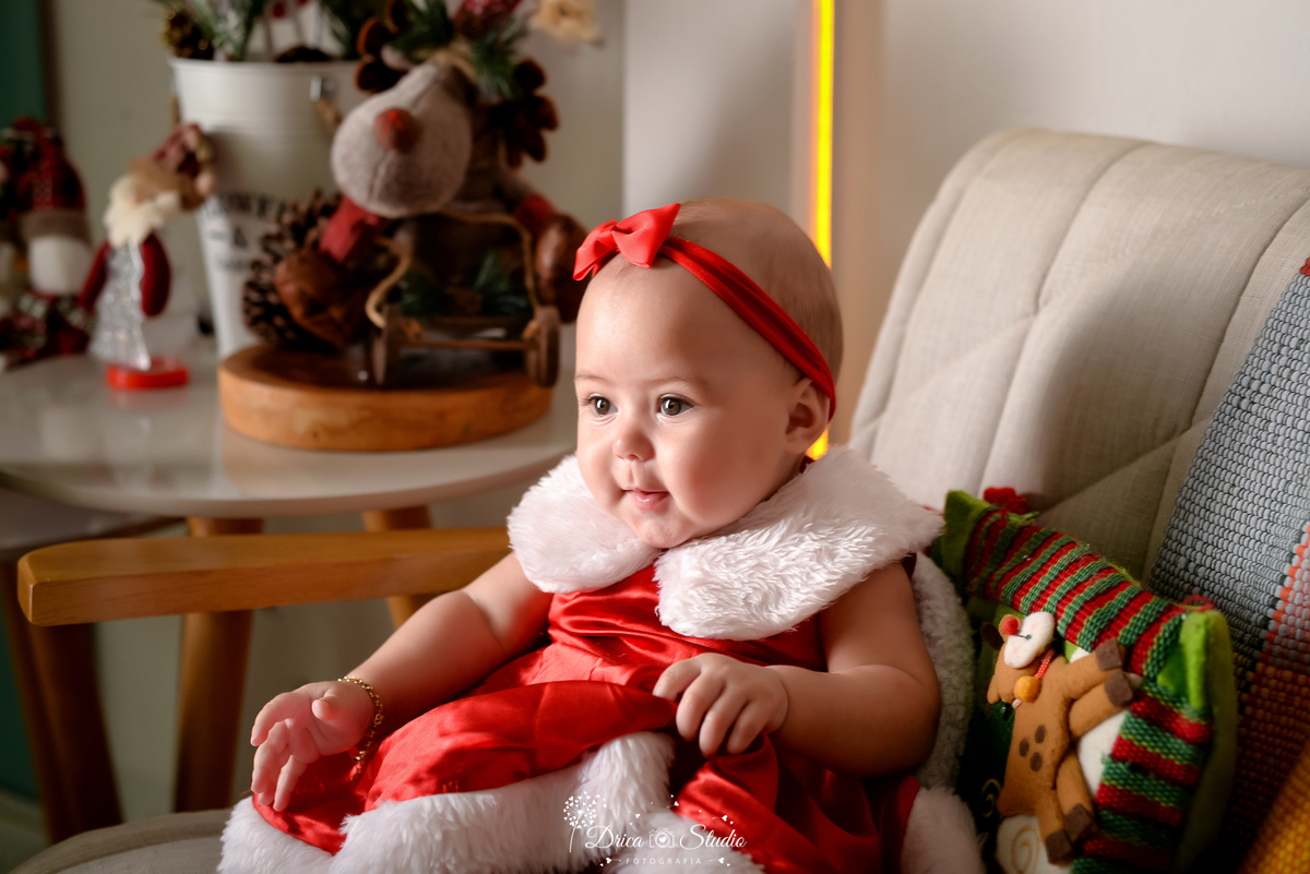 drica studio - ensaio fotografico - xinguara - fotos de natal  - bebê sentanda em poltroana vestindo roupinha de mamãe noel - rena de pelucia - almofada colorida - tiara vermelho - led amarelo - fundo branco