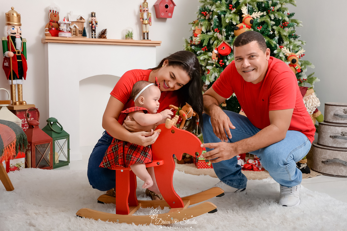 drica studio - ensaio fotografico - xinguara - fotos de natal - casal segurando bebê sentada em rena vermelha de madeira - casal veste camisa vermelha e calça jeans - árvore de natal - lareira branca - quebra nozes grande - lambião vermelho e verde 
