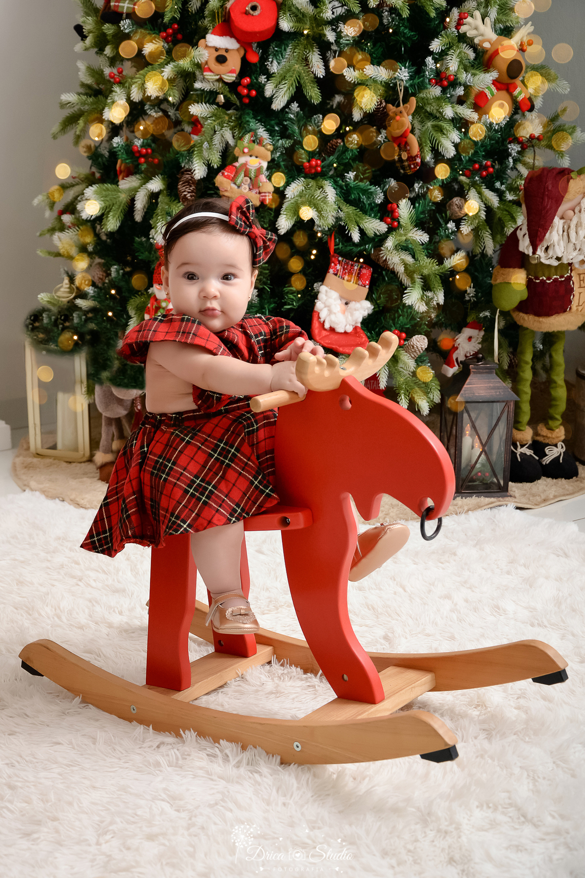 drica studio - ensaio fotografico - xinguara - fotos de natal - bebê usando vestido xadres vermelho com preto, sentada em rena vermelha de madeira - lampiões - papai noel de pelucia - árvore de natal - tapete felpudo branco
