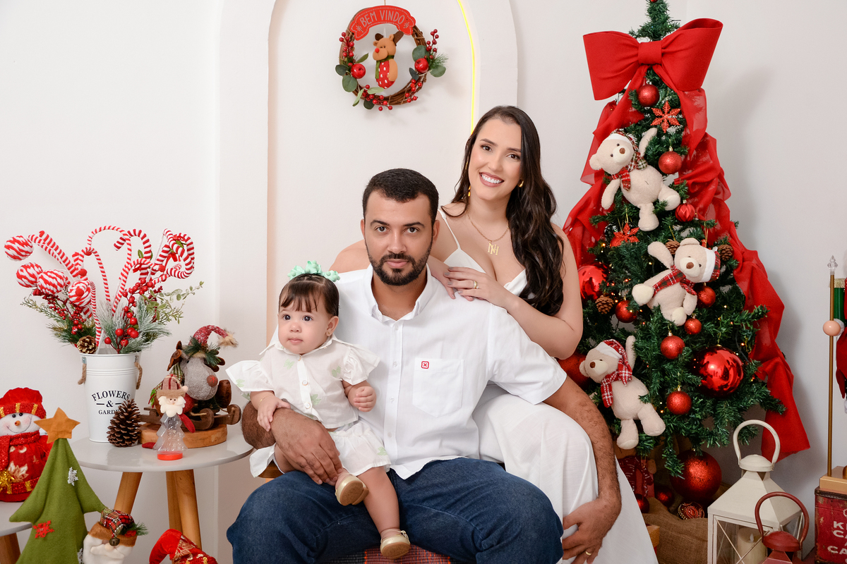 drica studio - ensaio fotográfico - xinguara - fotos de natal - casal sentado em poltrona com bebê no colo do pai, homem usa camisa branca e calça jeans, mulhere bebê bestido branco - decoração de natal - árvore de natal - lampiões - usrinhos natalinos