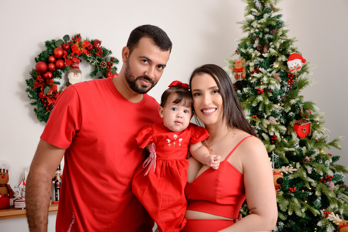 drica studio - ensaio fotográfico - fotos em família - xinguara - Pará - casa segurando filho nos braços - mulher veste croped vermelho, bebê veste vestido vermelho e homem veste camisa vermelha - decoração de natal - árvore de natal - guirlanda