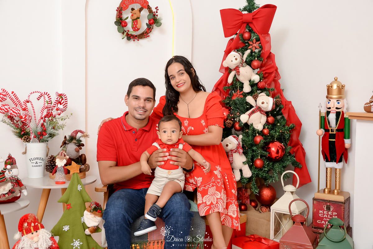 drica studio - ensaio fotografico - xinguara - fotos de natal - família pousa para foto de natal - homem veste camisa vermelha e calça jeans, bebê veste camisa vermeha e short bege, mulher veste vestido vermelho com detalhes branco - decoração de natal - 