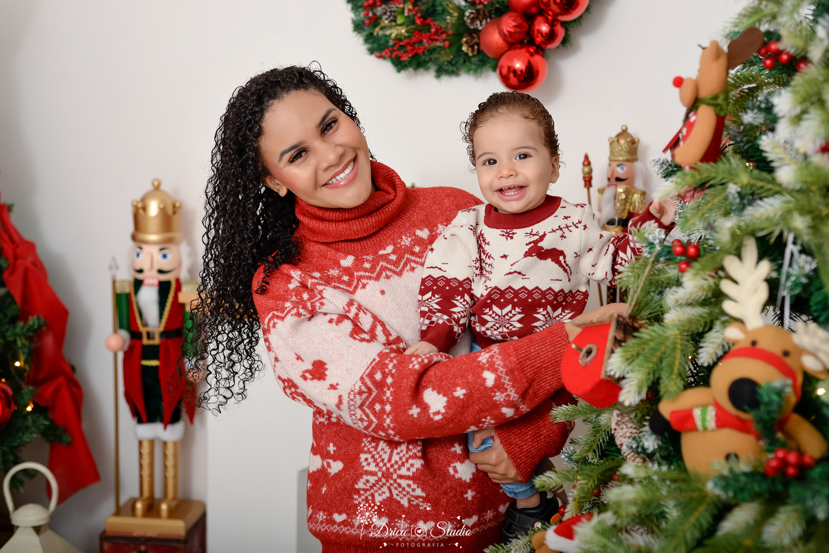 drica studio - ensaio fotografico - xinguara - fotos de natal - mãe vestindo sueter natalino segurando filho nos braças, que também veste sueter natalino - árvore de natal - quebra nozes - lampiões - fundo branco