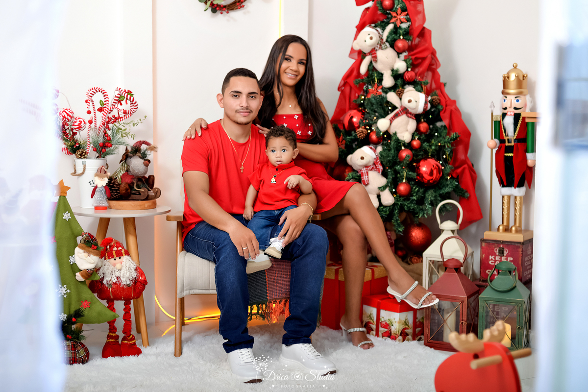drica studio - ensaio fotografico - xinguara - fotos de natal - pai sentado em poltono, esposa senta no braço da poltrona e bebê sentado no colo do pai - árvore de natal - quebra nozes grande - lampiões - papai noel de pelucia - laço vermelho sobre árvore