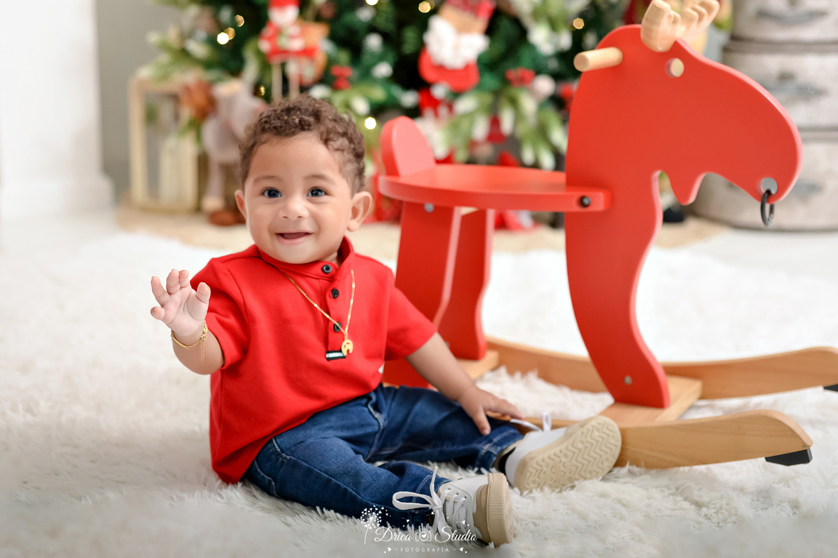 drica studio - ensaio fotografico - xinguara - fotos de natal - bebê sentado no carpete branco ao lado de rena vermelha de madeira - árvore de natal - presentes - bebê veste camisa vermelh e calça jean e calça tênis branco