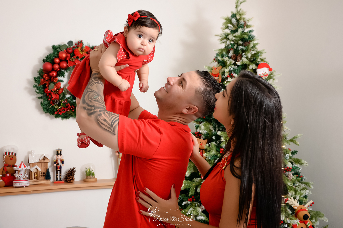 drica studio - ensaio fotografico - xinguara - fotos de natal - homem segurando filha eleva ao ar, veste camisa vermelha e bebê vestido vermelho com laço vermelho, mãe veste blusa vermelha - decoração de natal - árvore de natal - fundo branco