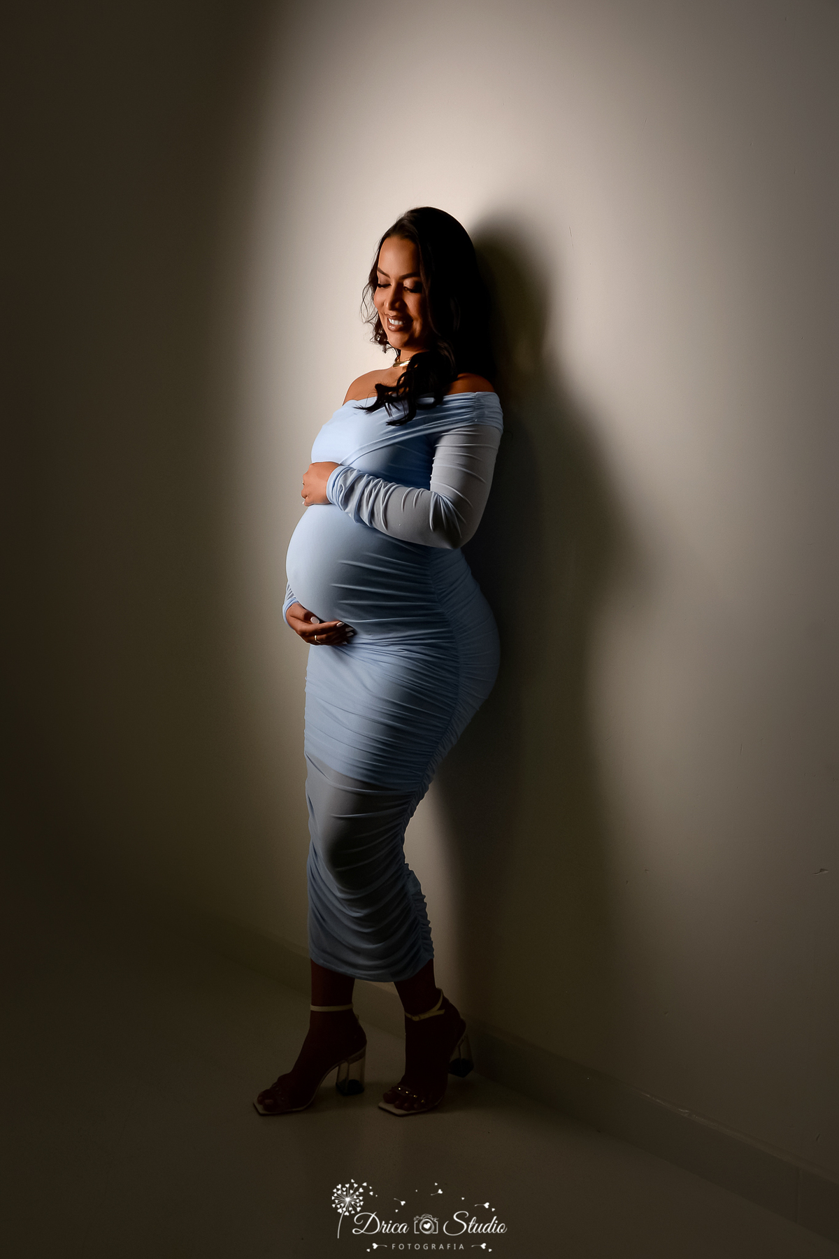drica studio fotográfico - ensaio gestante - xinguara - pará - gestante usando vestido longo e colado na cor azul claro - encostada em parede, coma a mãos envolta da barriga- meia luz dando um contrate com sombras - fundo neutro