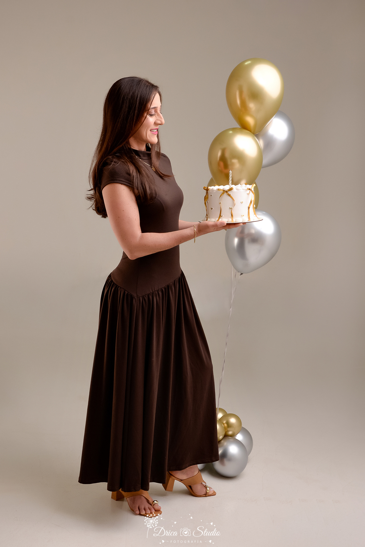 drica studio fotográfico - ensaio aniversário 40 anos - xinguara - pará - mulher usando vestido longo marrom, segurando bolo branco decorado com fita de cetim - balões dourado e prateado - fundo neutro