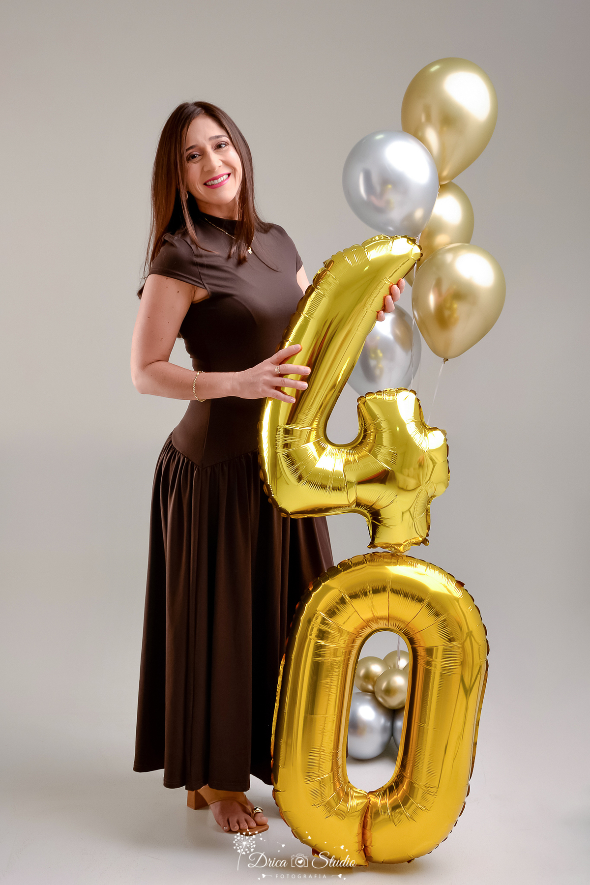 drica studio fotográfico - ensaio aniversário 40 anos - xinguara - pará - mullher usando vestido longo marrom, segurando numero dourado - sandalia de salto - fundo neutro