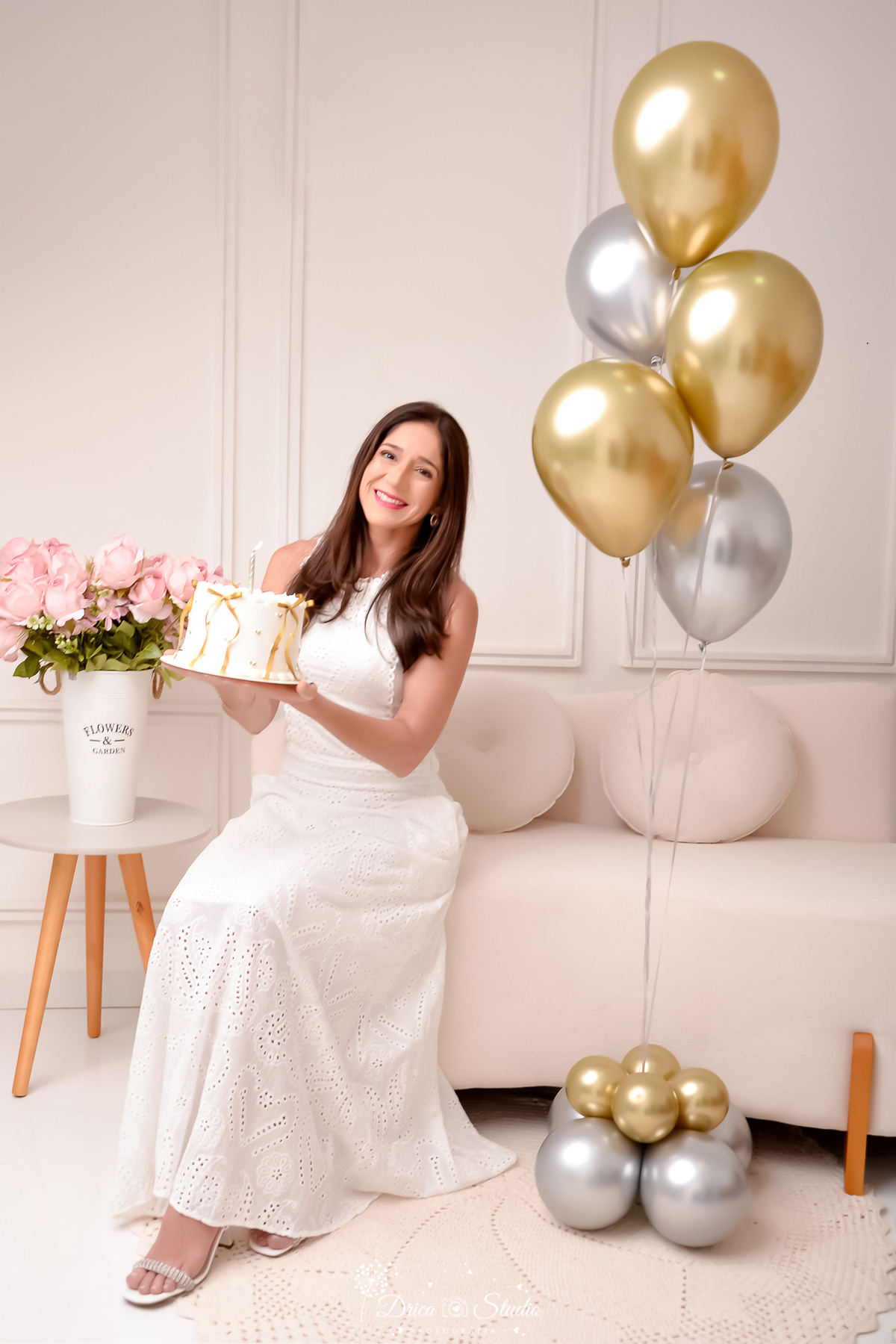 drica studio fotográfico - ensaio aniversário 40 anos - xinguara - pará - mulher usando vestido branco longo, segurando bolo branco decorado com fitas de cetim douradas - balões dourados e prateado - sofá bege claro - vaso com rosas - mesinhas de canto