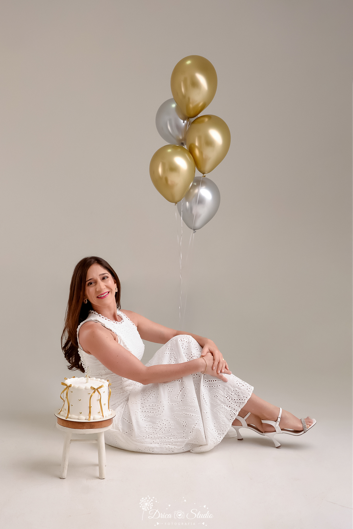 drica studio fotográfico - ensaio aniversário 40 anos - xinguara - pará - mulher sentada na chão com mão en volta das pernas - bolo branco decorado com fitas de cetim - balões dourado e prateado - fundo neutro