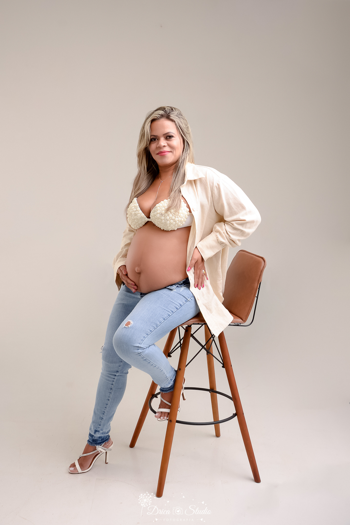 drica studio fotográfico - ensaio gestante - xinguara - pará - mulher grávida sentada em banqueta, usando jeans, camisa clara e top delicado, em fundo neutro