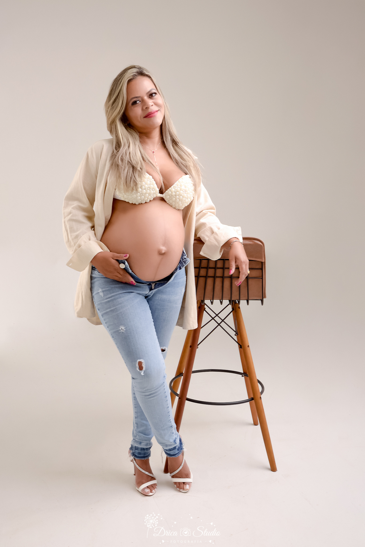 drica studio fotográfico - ensaio gestante - xinguara - pará - mulher gravída em pé apoida em banqueta de couro e madeira, usando camisa clara e top delicado, em fundo neutro