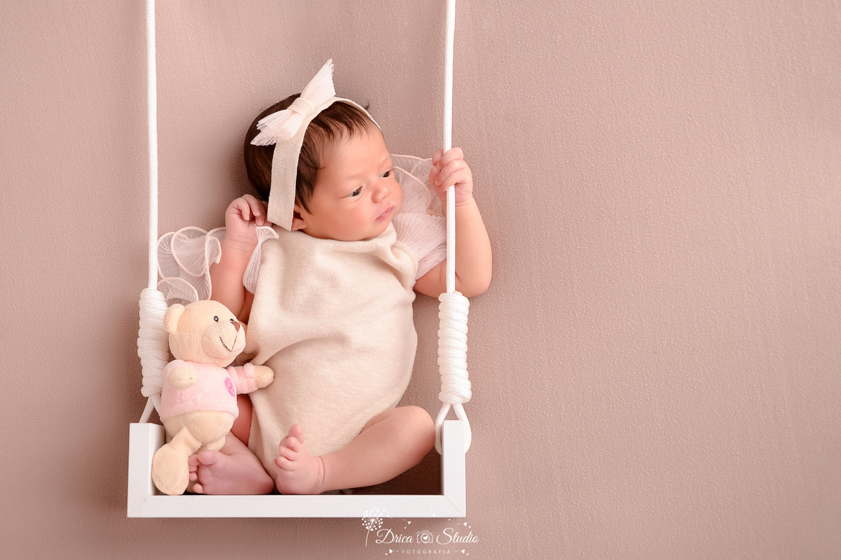 drica studio fotográfico - ensaio newborn - xinguara - pará - bebê recém-nascido sentada em balanço suspenso, sobre um fundo bege -bebê veste roupinha clara e delicada, com detalhe em tule, faixa de cabelo com laço e segura um ursinho de pelúcia