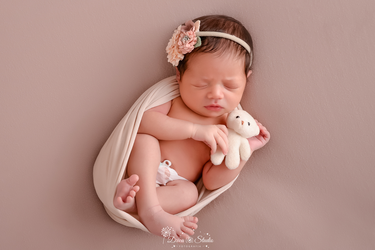 drica studio fotográfico - ensaio newborn - xinguara - bebê recém-nascido envolvido em tecido claro, deitada com as pés cruzados, segurando um ursinho de pelúcia claro - 