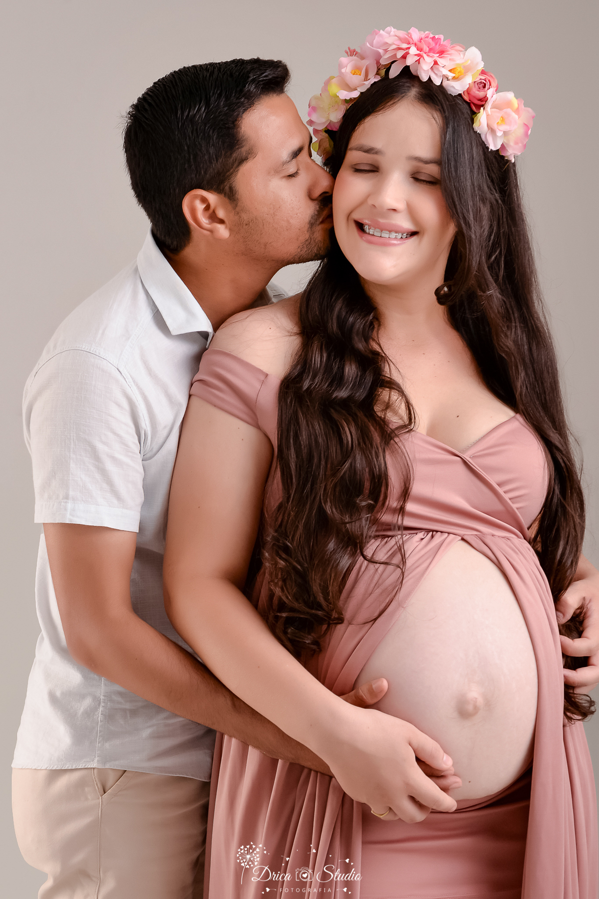 drica studio - ensaio fotográfico gestante - xinguara - pará - gestante usando vestido ombro a ombro rose com abertura frontal deixando a barriga a mostra, esposo a envolve com os braços e beija suavemente seu rosto - fundo neutro