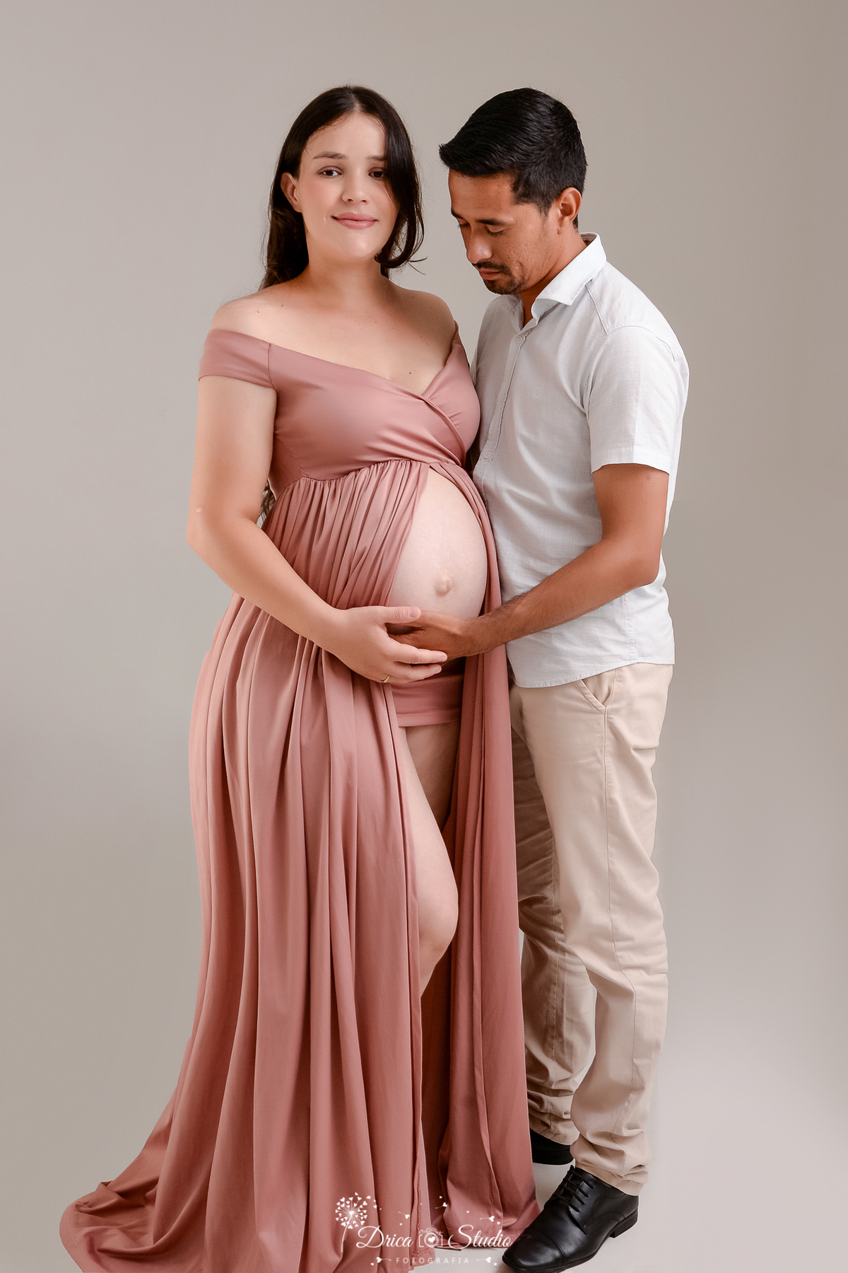 drica studio - ensaio fotográfico gestante - xinguara - pará - casal posam para foto de gestante, ela usa vestido longo rose aberto na frente deixando a barriga a amostra  e  esposo veste camisa branca e calça bege - fundo neutro