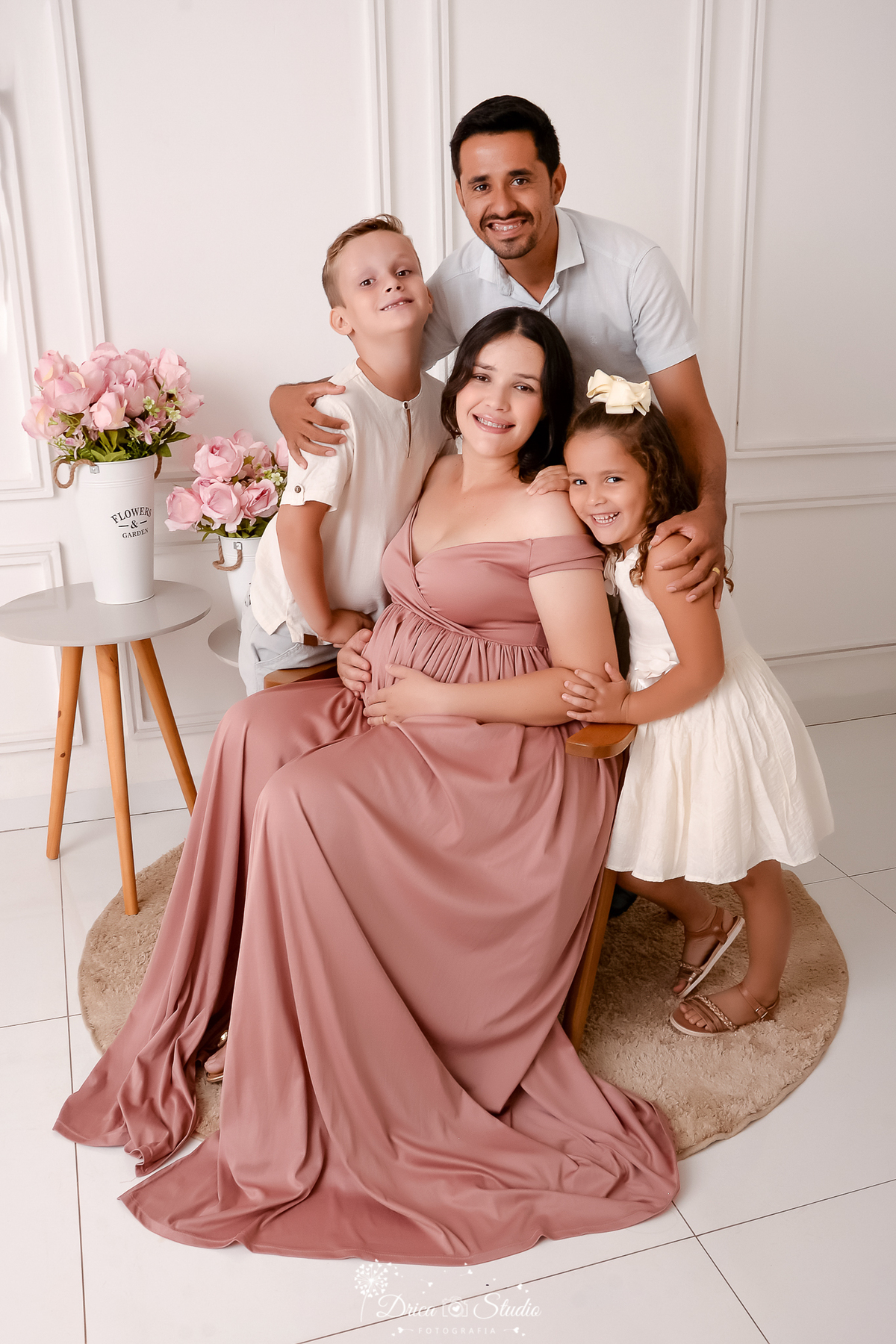 drica studio - ensaio fotográfico gestante - xinguara - pará - Família em ensaio de gestação em estúdio, mãe grávida sentada com vestido rosé, pai e filhos abraçados, sorrisos naturais, cenário clean com flores, fotografia familiar delicada e profissional
