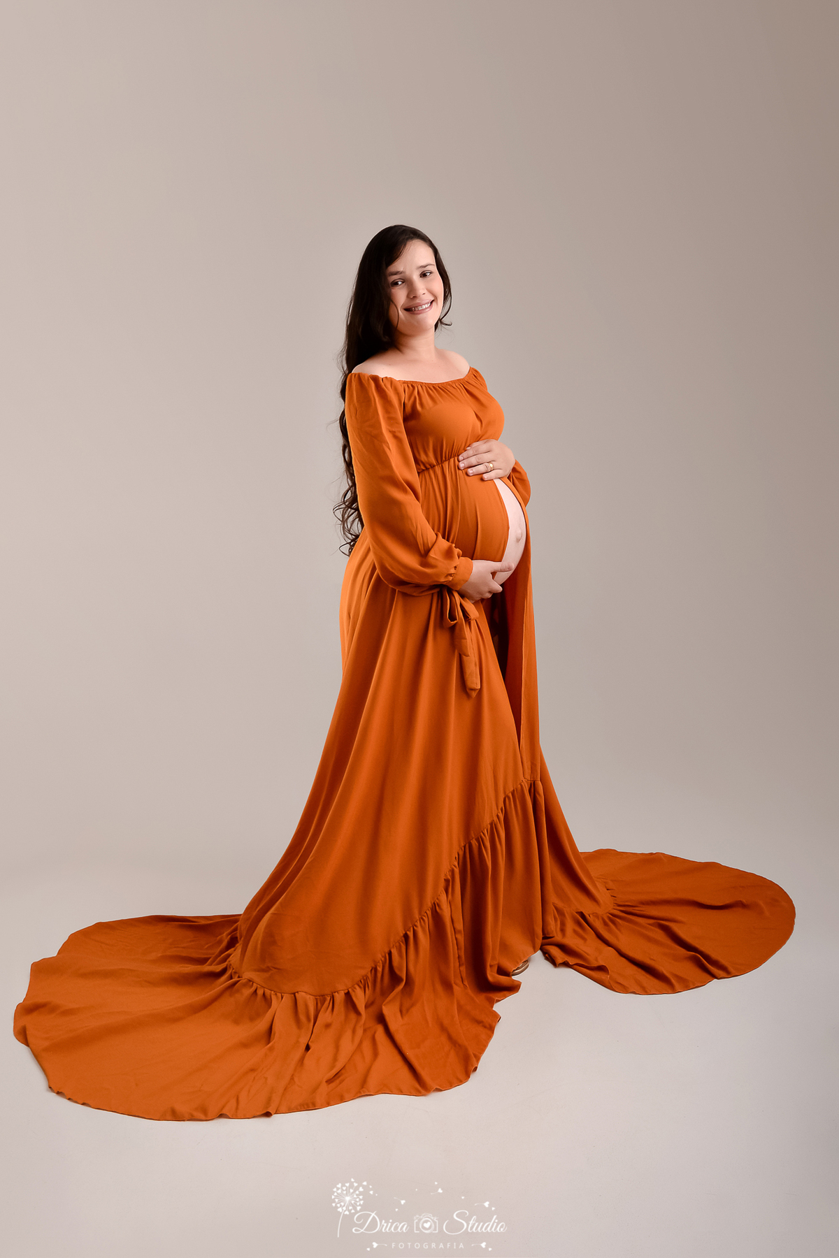 drica studio - ensaio fotográfico gestante - xinguara - pará - Gestante em ensaio de gestante em estúdio, vestido longo terracota com cauda, mãos na barriga, sorriso suave, fundo neutro, fotografia elegante e profissional.