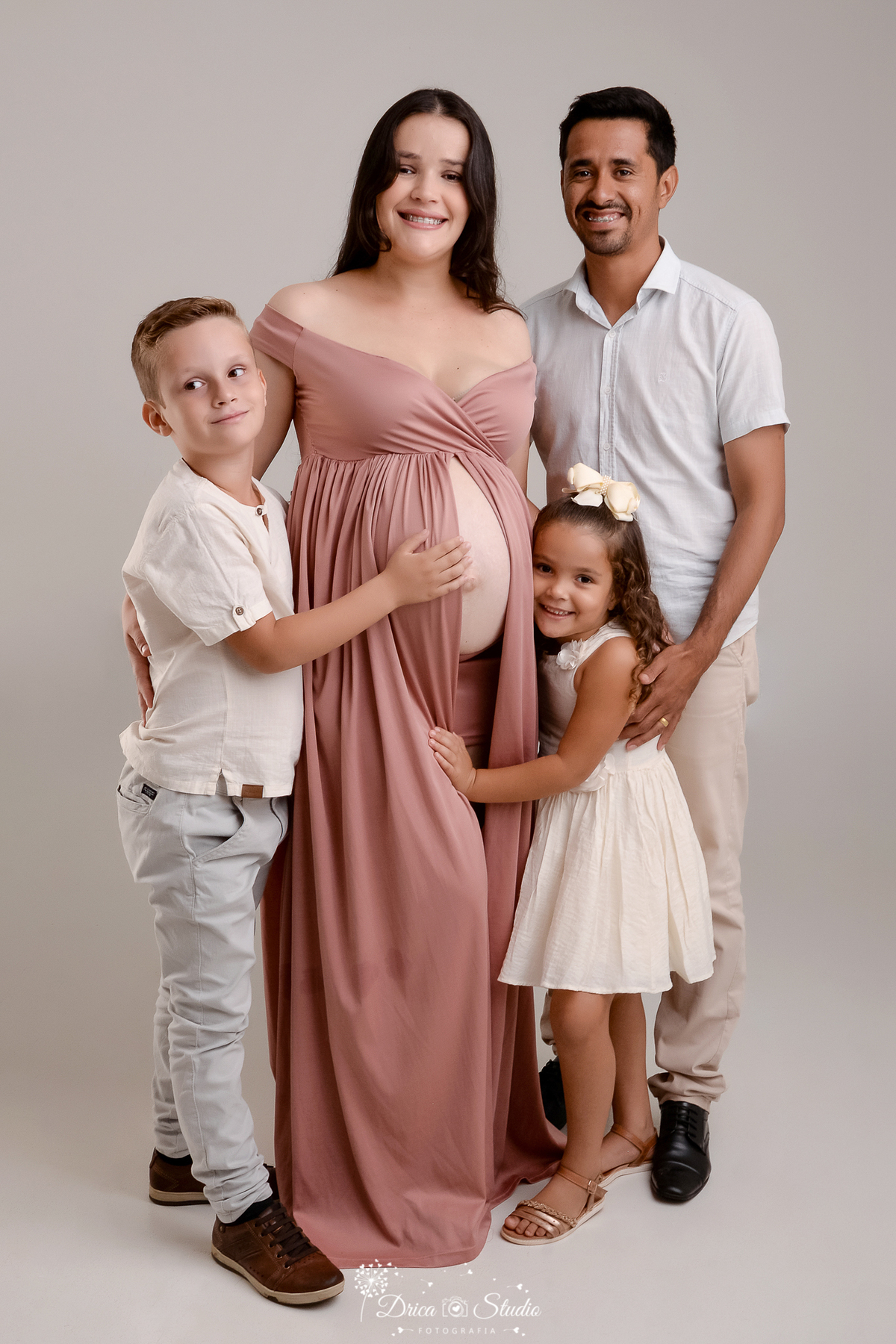 drica studio - ensaio fotográfico gestante - xinguara - pará - gestante fazendo foto com a familia, ela usa vestido longo rose, menina usa vestido bege claro, menino camisa bege e calça jeans, esposo caisa branca e calça bege - lfundo neutro