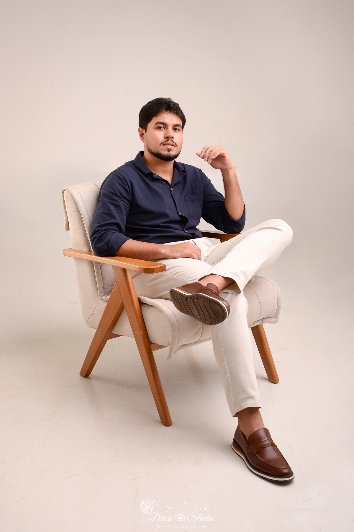 drica studio - ensaio fotografico formatura - administração - xinguara - Pá - rapaz sentado em poltrona clara com detalher me madeira, expressão seria- veste camisa azul marinho e calça bege clara - fundo clean