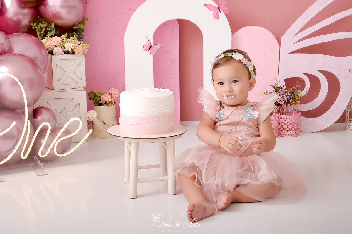 drica studio-ensaio infantil-smash the cake-jardim de borboletas-xinguara-pará-bebê sentada com bolo ao lado - one de led-caixotes branco-balões rosa- arco rosa- asa de borboleta- vestido rosa e tiara de flores - fundo rosa