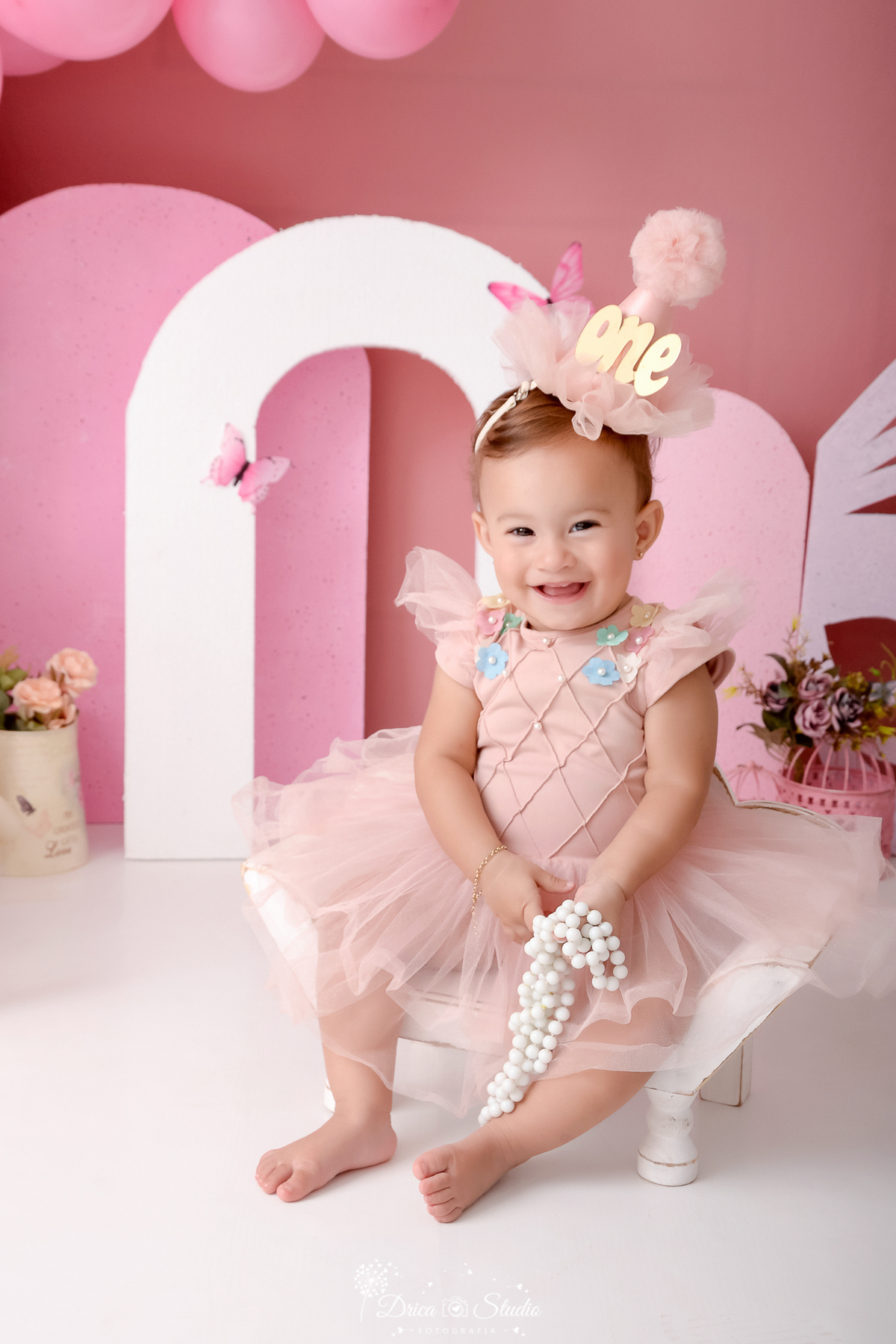 drica studio-ensaio infantil-smash the cake-jardim de borboletas-xinguara-pará-close da bebê sorrindo sentada-bebê sorrindo com vestido rosa e tiara de chapéuzinho com nome one em acrilico - flores rosa - asa de borboleta - fundo rosa