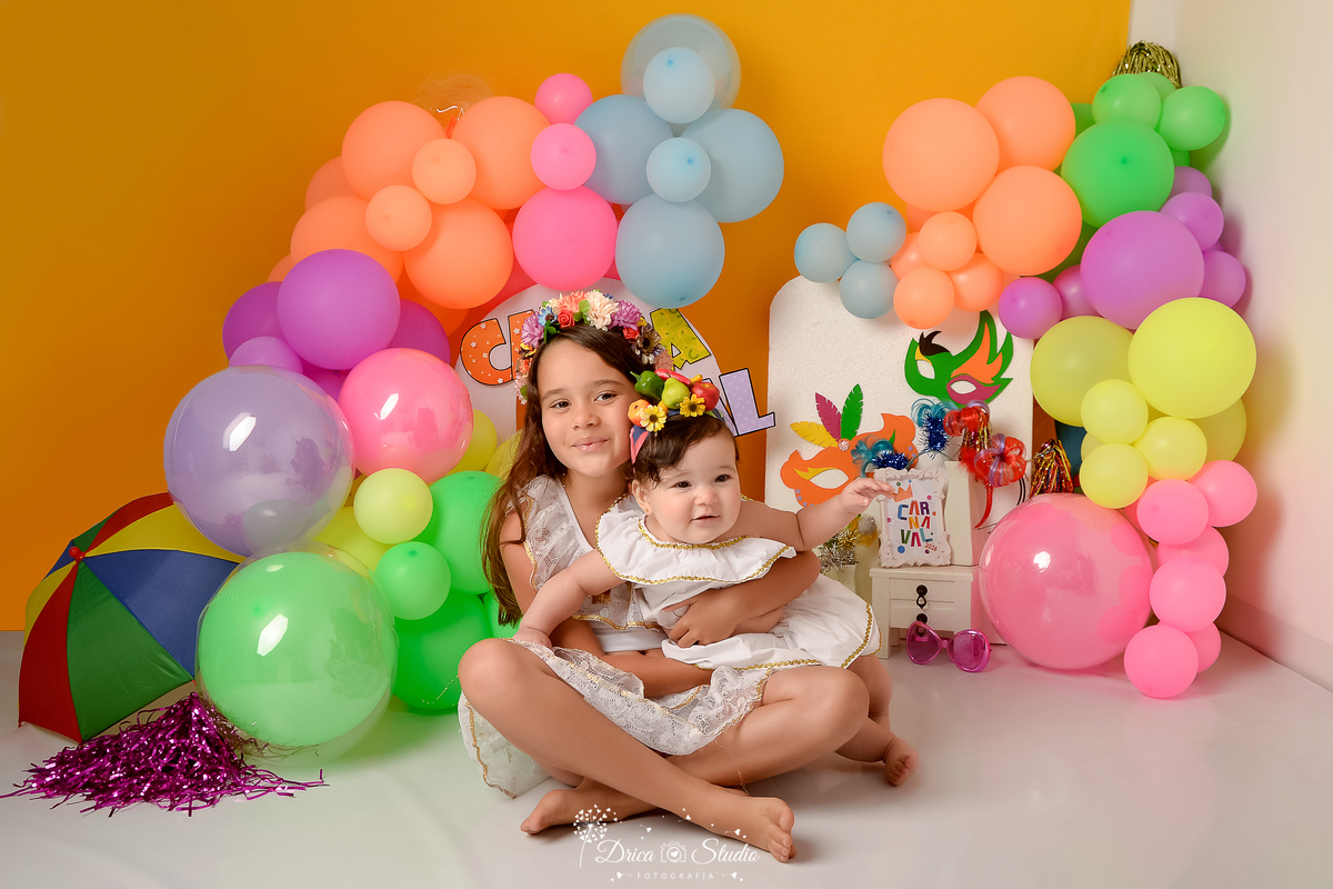 drica studio - ensaio fotográfico infantil - carnaval- xinguara - pará - balões coloridos - sombrinha de frevo - máscaras de carnaval - bebê vestida de carme miranda no colo da irmã - penteadeira branca - painel branco e lilas- pompom rosa escuro
