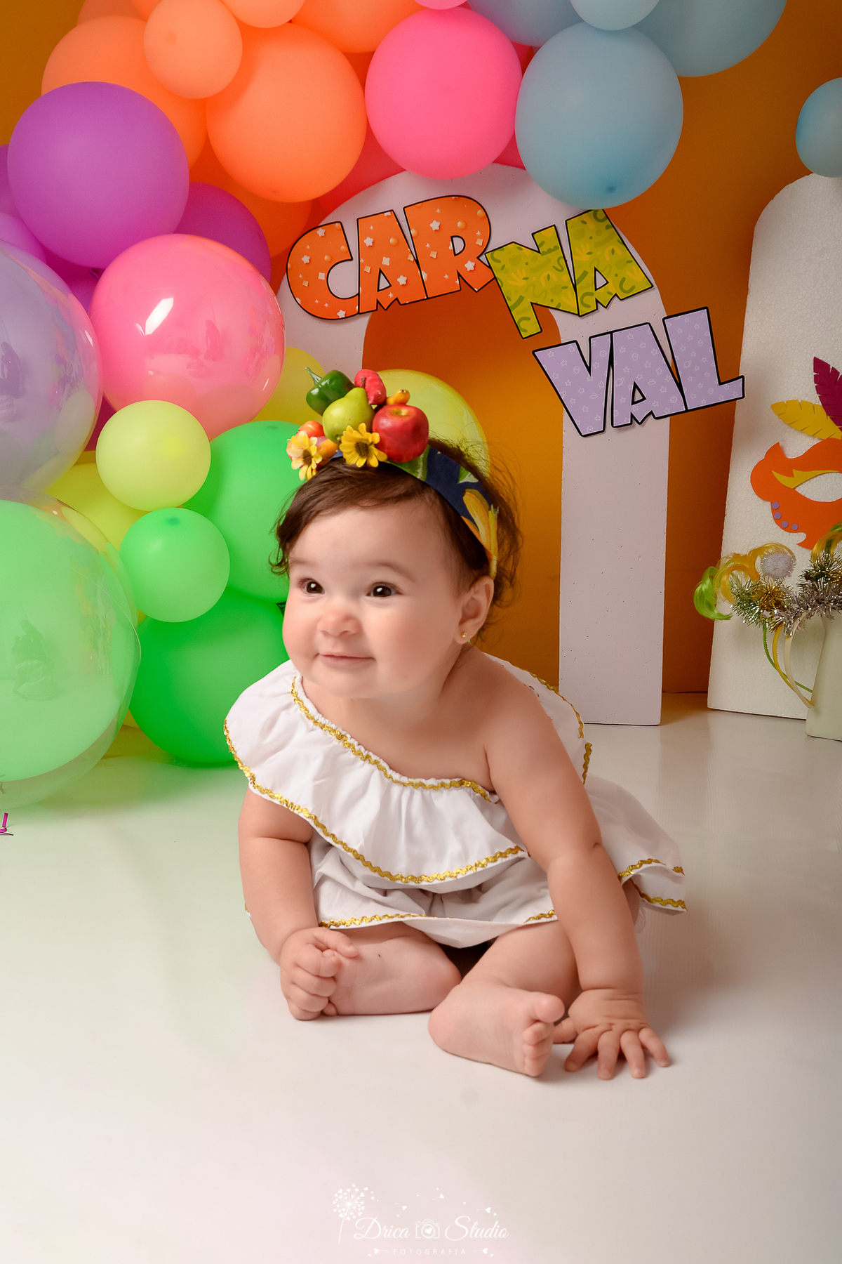 drica studio - ensaio fotográfico infantil - carnaval- xinguara - pará - bebê sentada no chão vestida de carmem miranda, com frutas artificiais na cabeça - balões coloridos - painel branco e lilar - máscara de carnaval - fundo amarelo