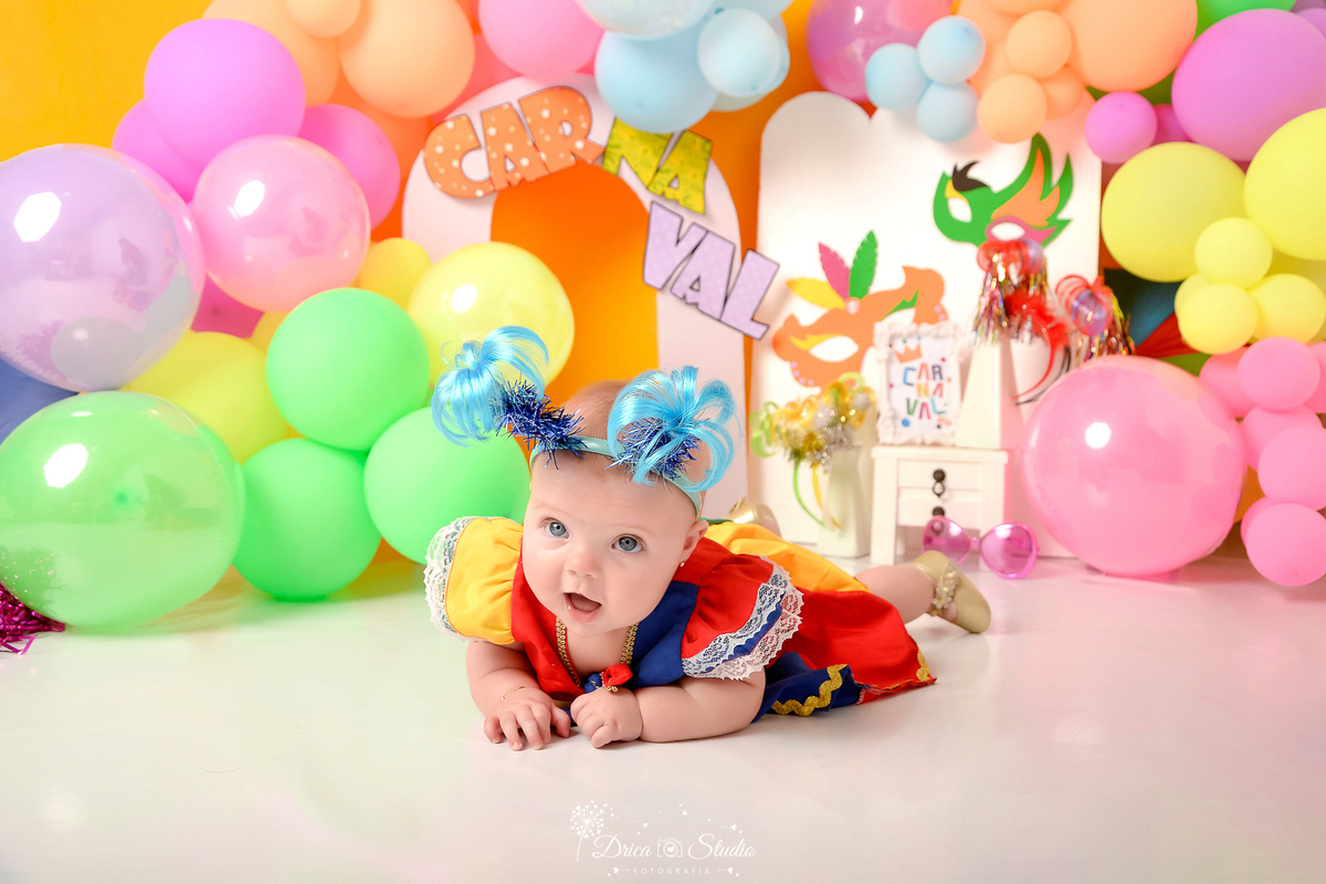 drica studio - ensaio fotográfico infantil - carnaval- xinguara - pará - bebê deitada no chão de bruços vestida com roupa de frevo e usando tiara de coqueiro azul - balões coloridos- máscara de carnaval - fundo amarelo