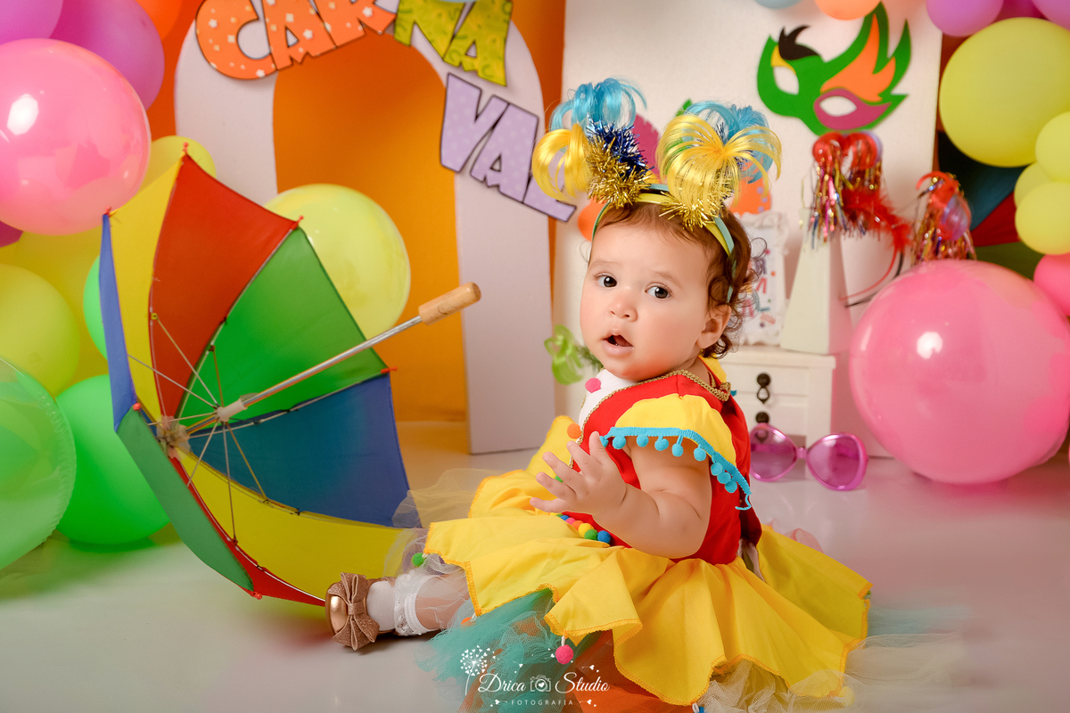 drica studio - ensaio fotográfico infantil - carnaval- xinguara - pará - bebê sentada no chão usano vestido de palhacinha e tiaras de coqueiro coloridas - sombrinha de frevo - balões coloridos - máscara de carnaval - penteadeira branca - fundo amarelo